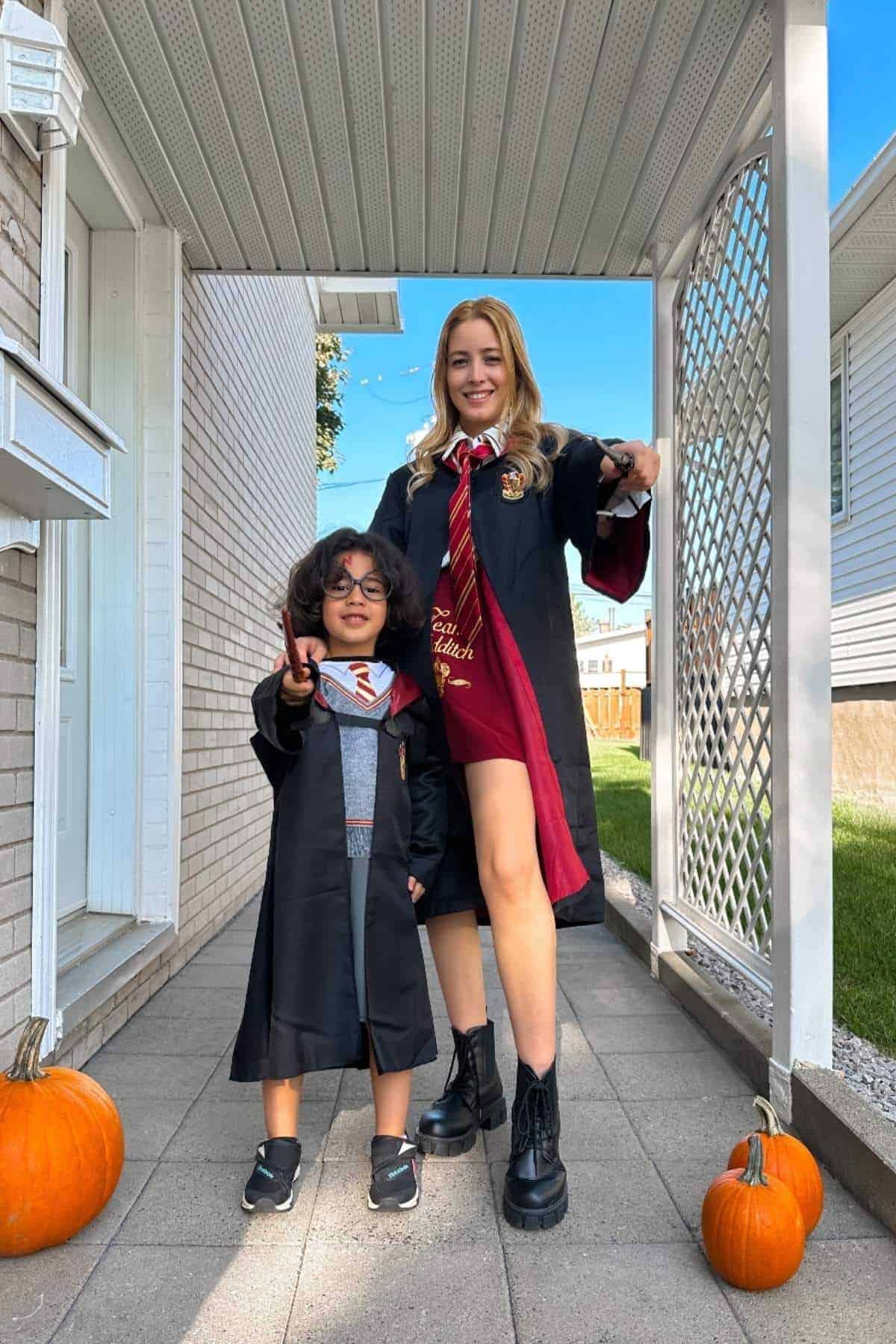 An adult and a child dressed in Hogwarts-inspired costumes stand on a porch with three pumpkins, holding wands and posing for a photo.