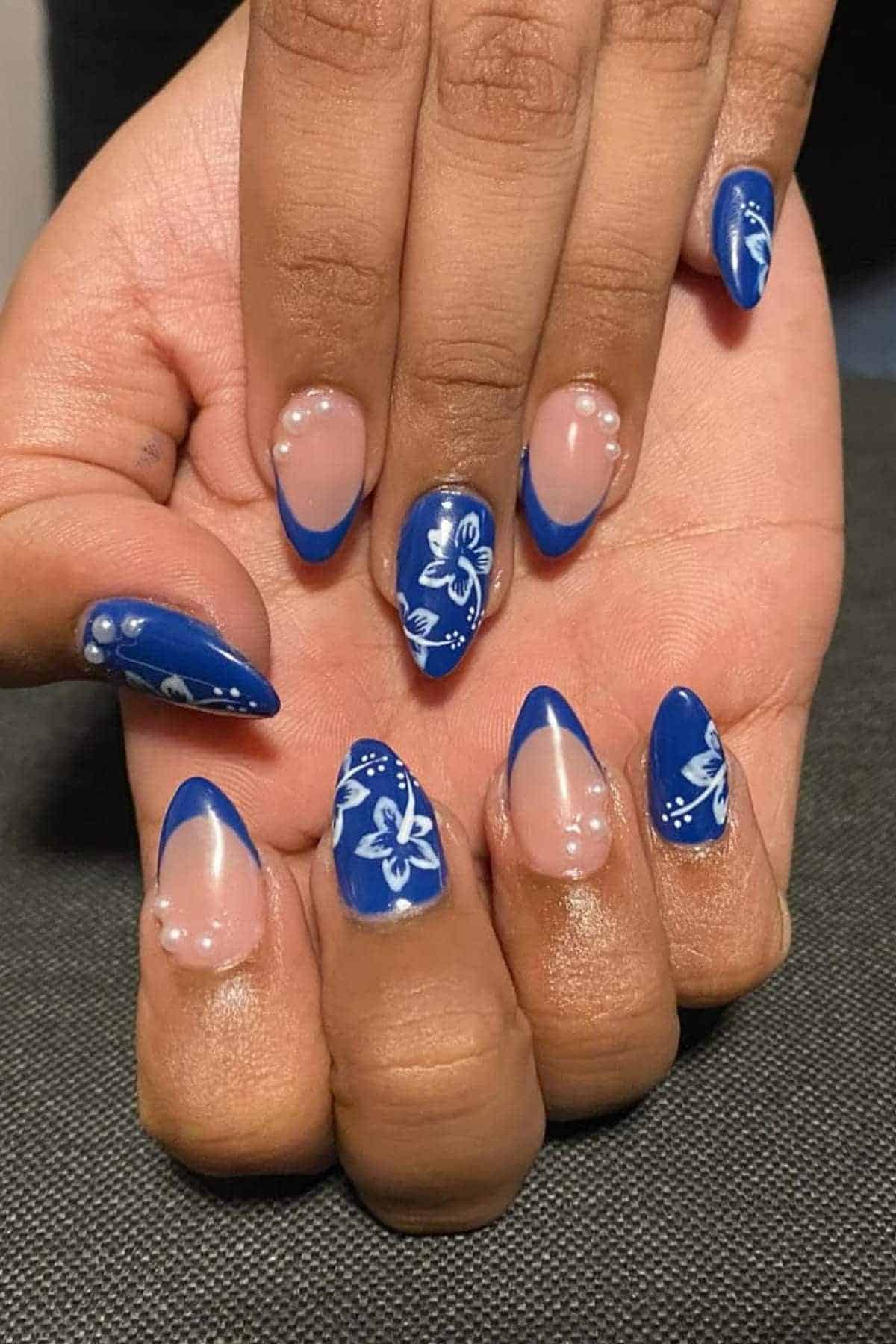 Hands with almond-shaped nails featuring blue French tips, white floral designs, and pearl accents on some nails, displayed against a dark background.