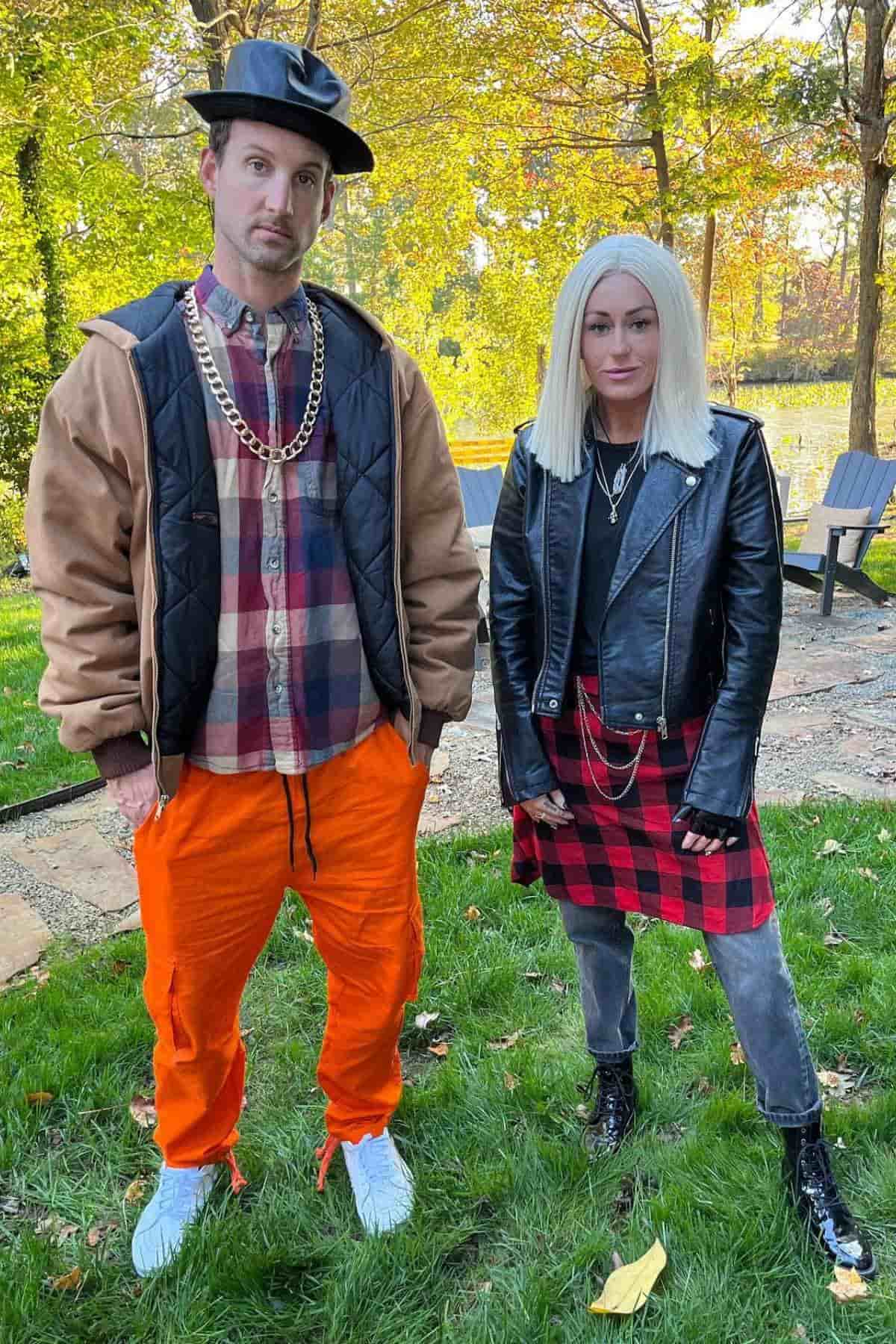 Two adults stand outdoors on grass. The man wears a hat, gold chains, plaid shirt, brown jacket, and orange pants. The woman wears a blonde wig, black jacket, plaid skirt, and gray jeans.