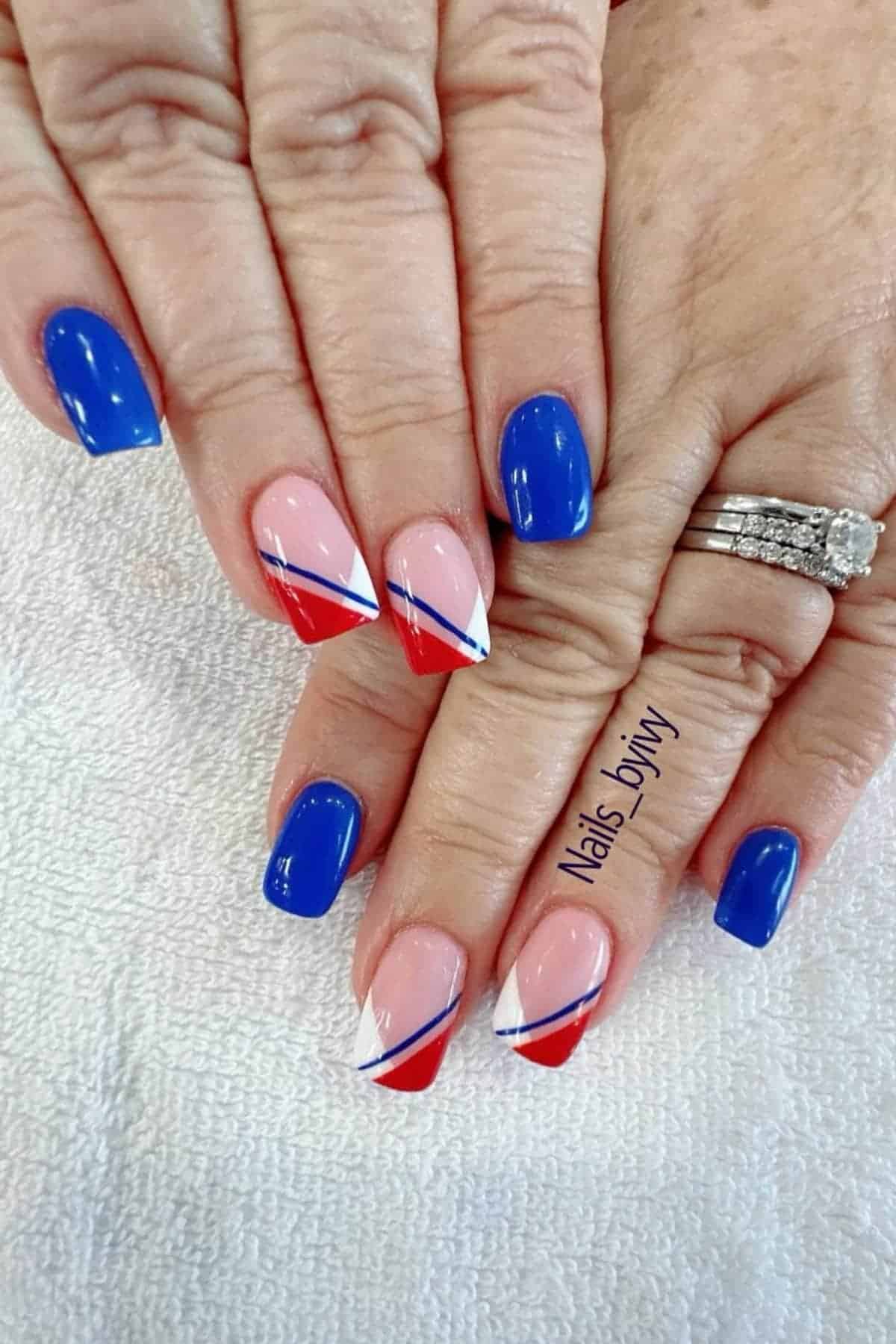 Hands with manicured nails featuring solid blue polish on some nails and red, white, and blue diagonal designs on others. The person is wearing rings on one hand.