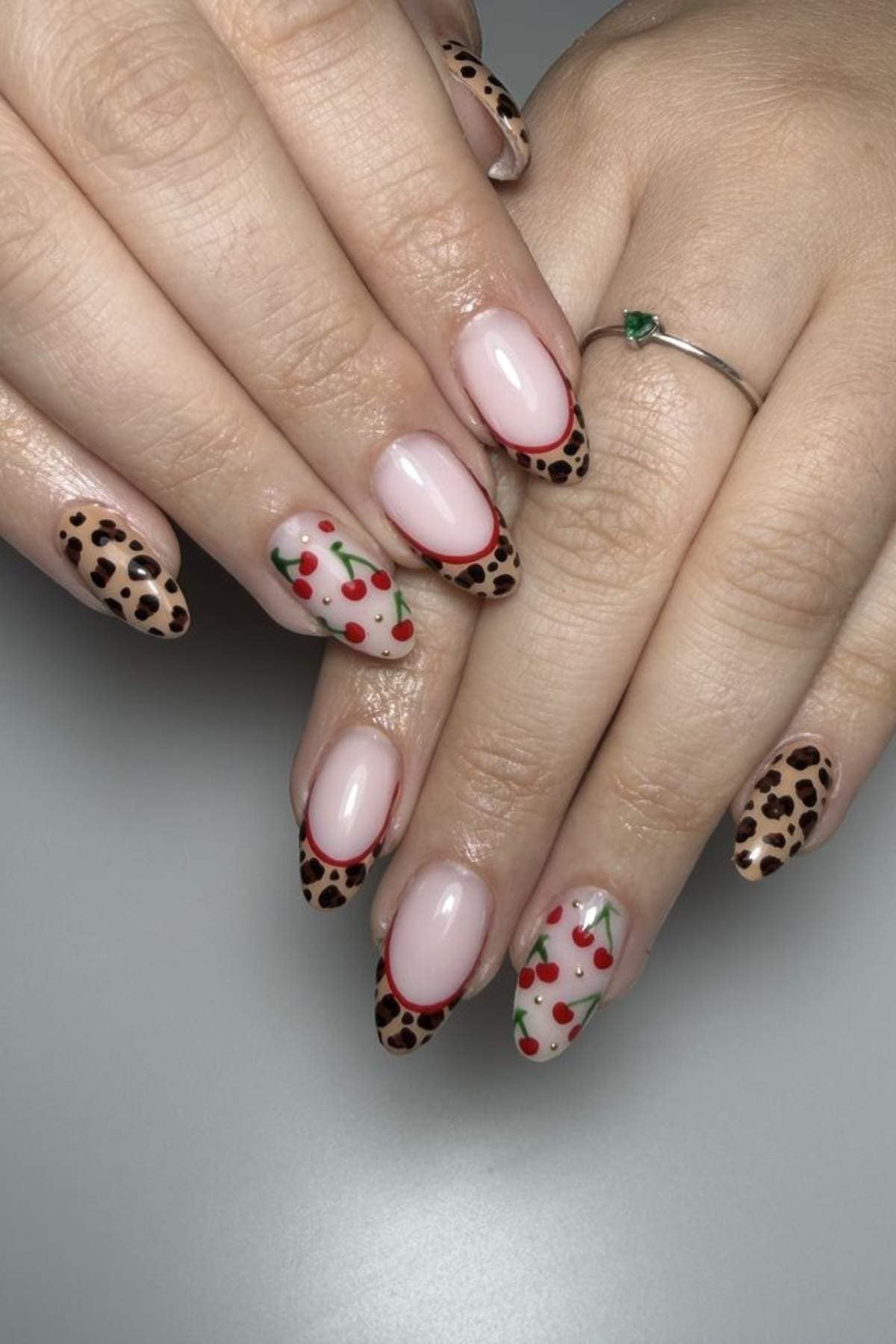 A hand with long almond-shaped nails features a mix of cheetah print and cherry designs on a light pink base. The person is wearing a thin silver ring with a small green stone.