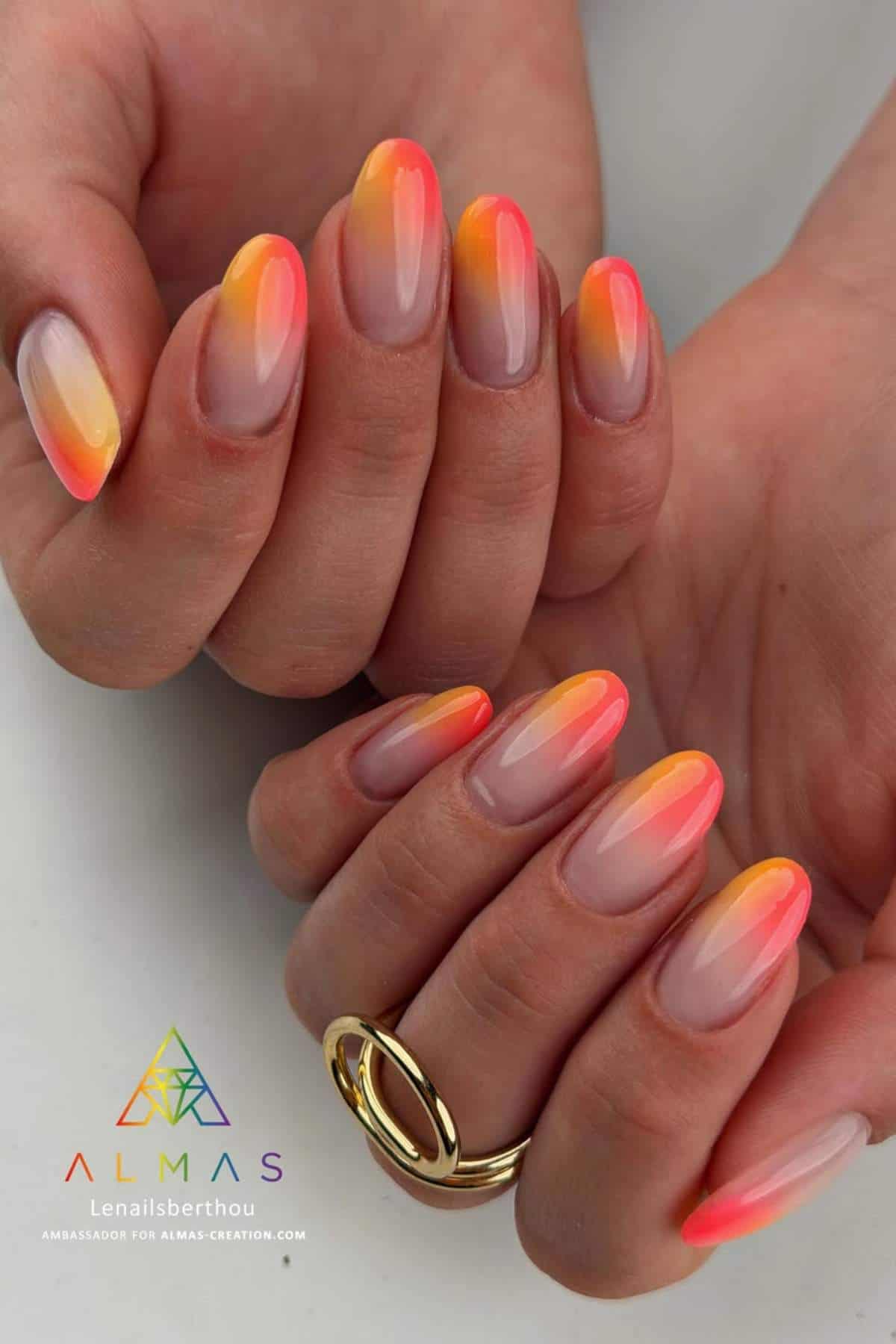 Close-up of hands with almond-shaped nails featuring a gradient design in pink, orange, and yellow shades. The person wears a gold ring on one finger. Almas branding visible at the bottom.