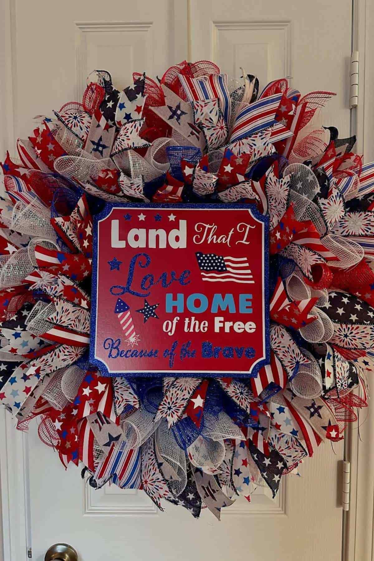 A patriotic wreath with red, white, and blue ribbons and stars, featuring a sign that reads "Land that I love, Home of the Free because of the Brave.