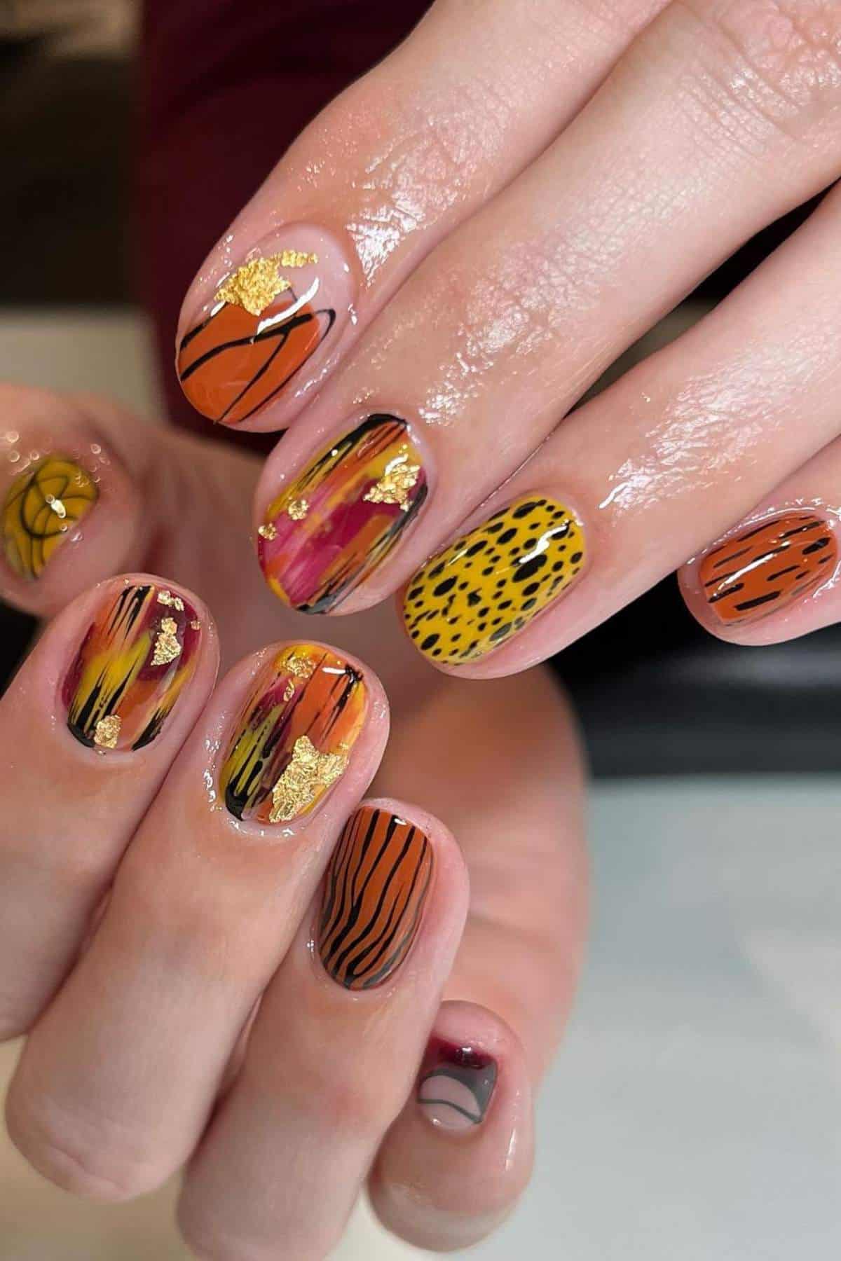 Close-up of hands with glossy, multicolored nail art featuring abstract animal prints, gold foil accents, and a mix of orange, yellow, and black patterns.