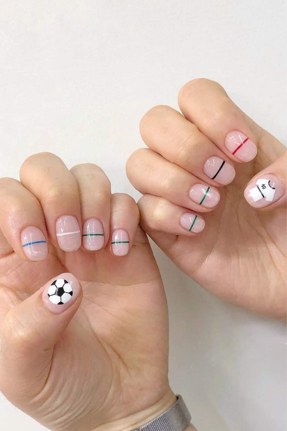 Hands with minimalist nail art featuring fine colored lines and a soccer ball design on one thumb against a plain white background.