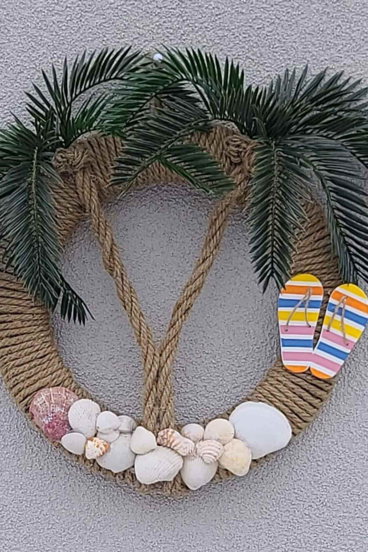 A decorative wreath made of rope, artificial palm leaves, seashells, and colorful flip-flops hangs on a gray wall.