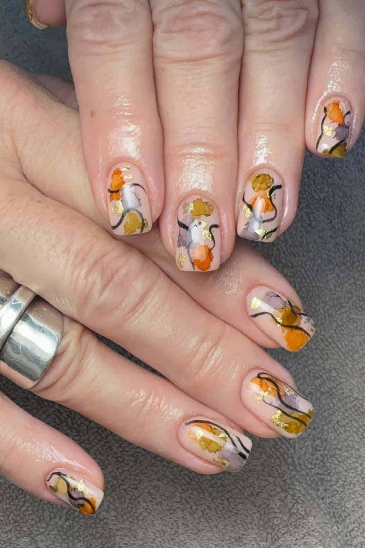 Hands with short nails featuring abstract floral nail art in orange, yellow, and brown tones on a neutral base. One hand wears a silver ring. Nails rest on a gray surface.