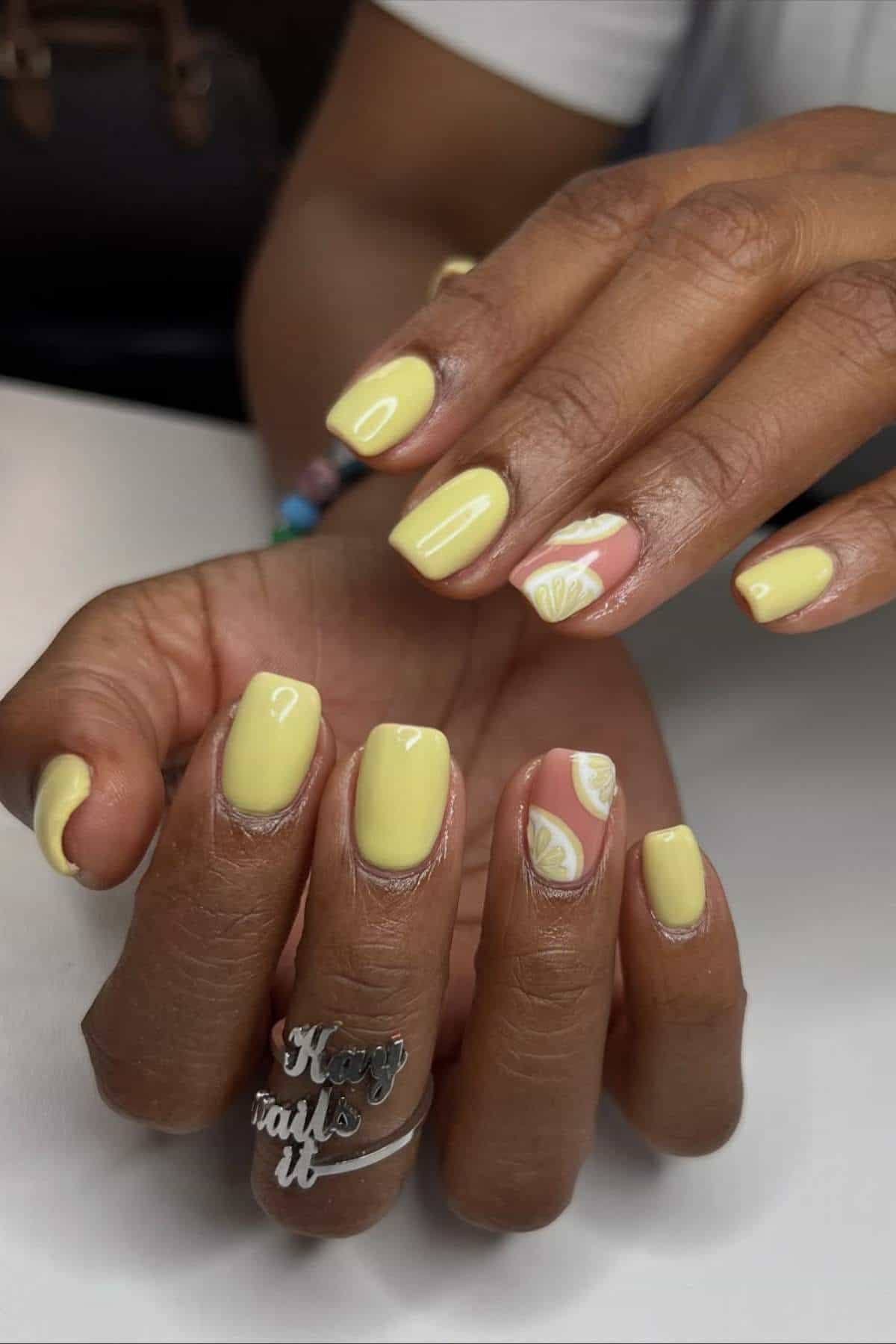 Short nails with light yellow polish. Two accent nails feature a pink background with white and yellow lemon slices.