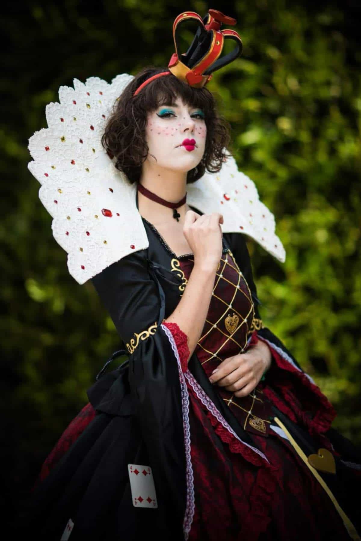 Person dressed in a Queen of Hearts costume with a large white collar, heart-shaped crown, and playing card accessories, standing outdoors against a green blurred background.