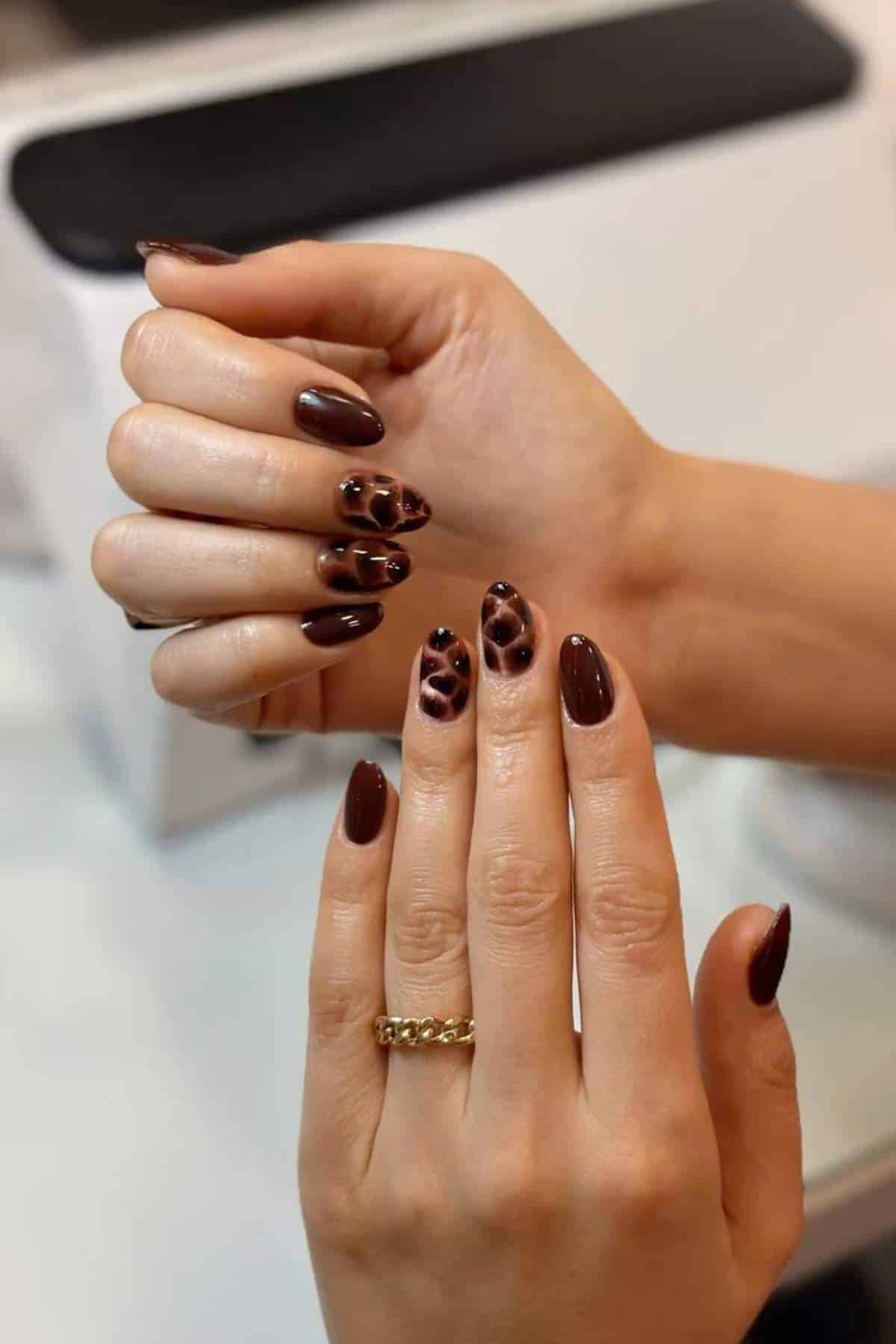 Hands with almond-shaped nails painted dark brown; ring fingers and middle fingers feature a leopard print design. A gold chain ring is worn on one finger.
