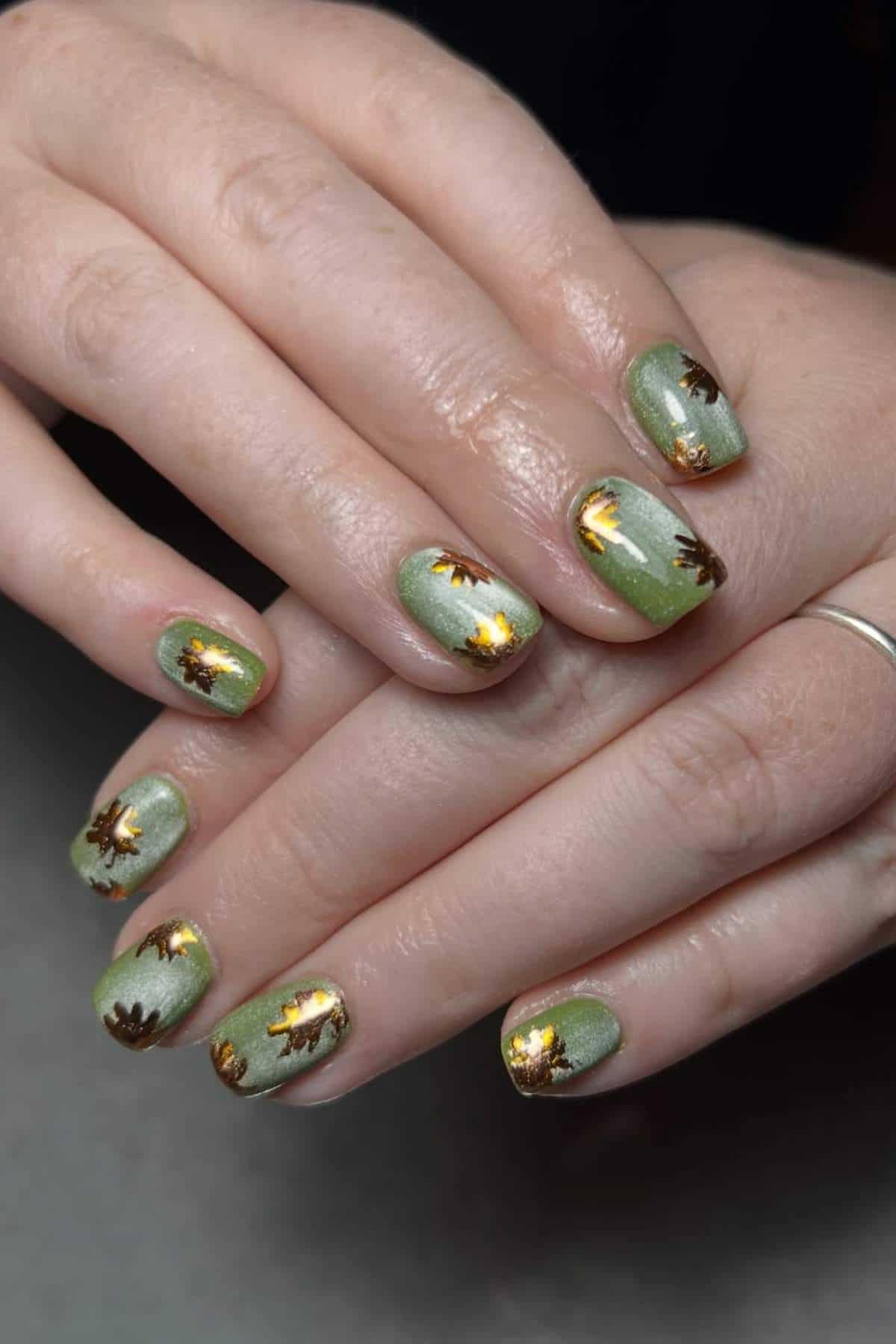 Hands with short, square-shaped nails painted green and decorated with gold and brown floral designs.