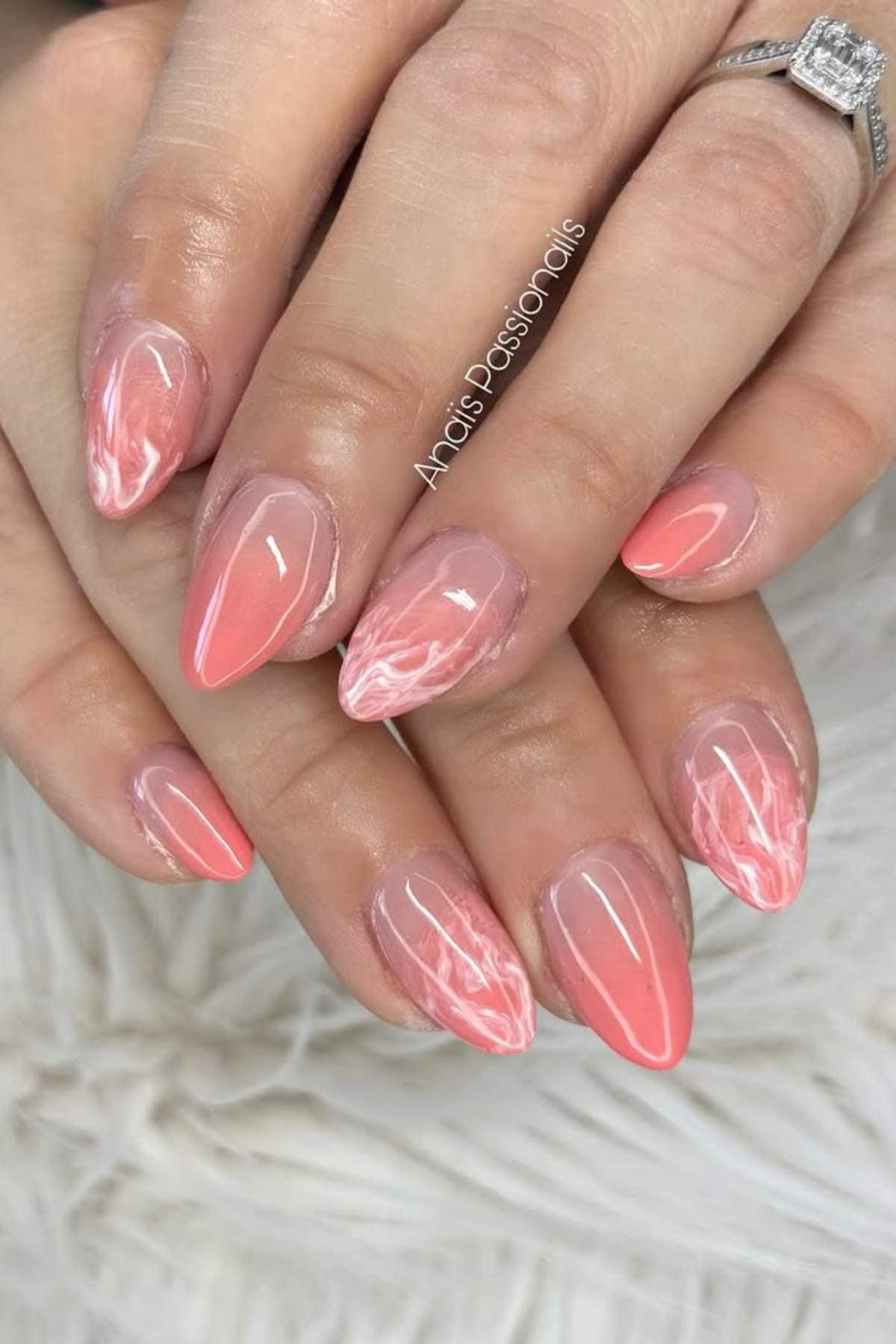 Hands with almond-shaped nails featuring a pale coral and white marble design on a soft background. One hand wears a silver ring with a square center stone.