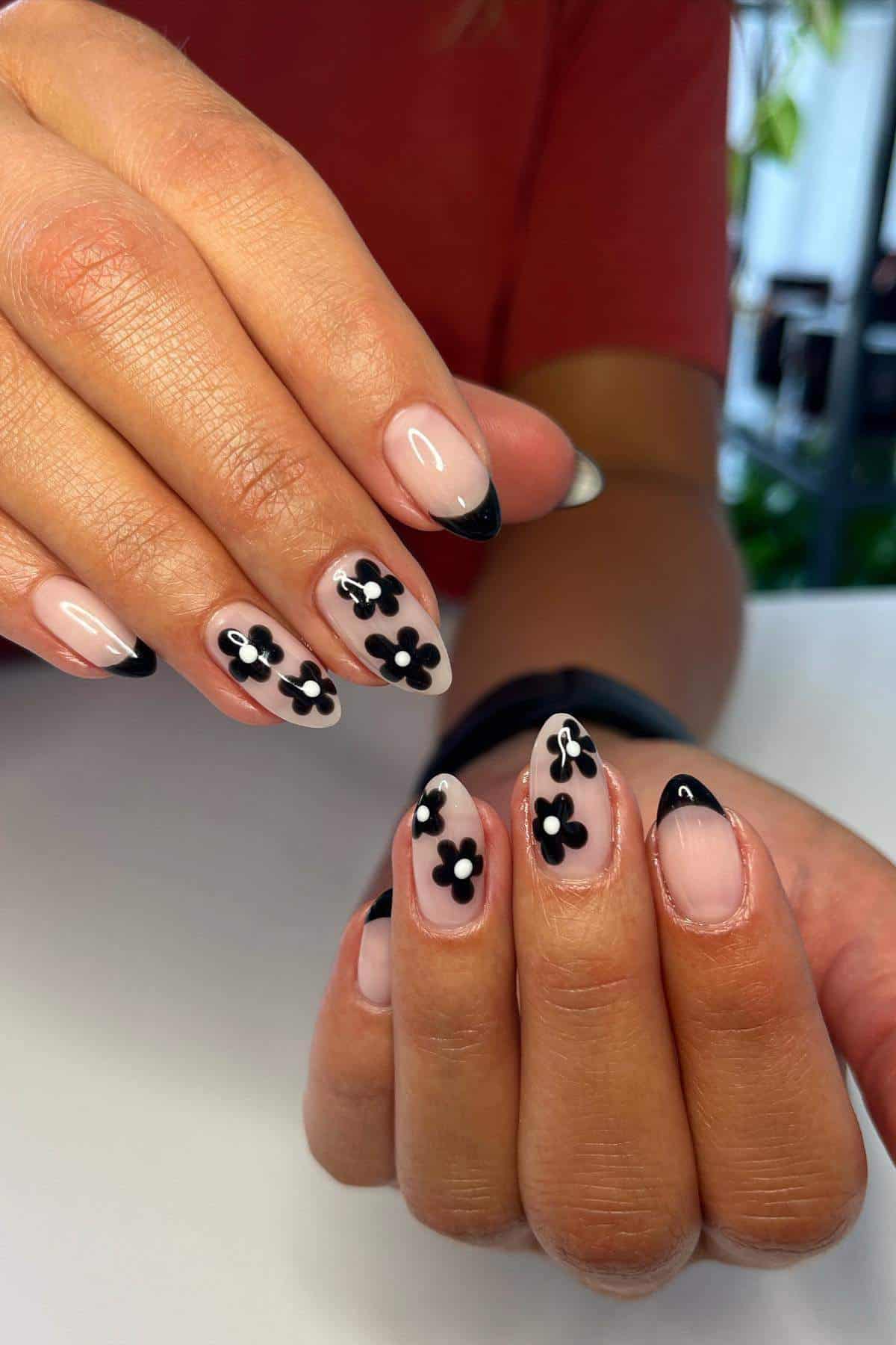 Hands with almond-shaped nails featuring a nude base, black French tips, and black and white flower nail art on each nail.