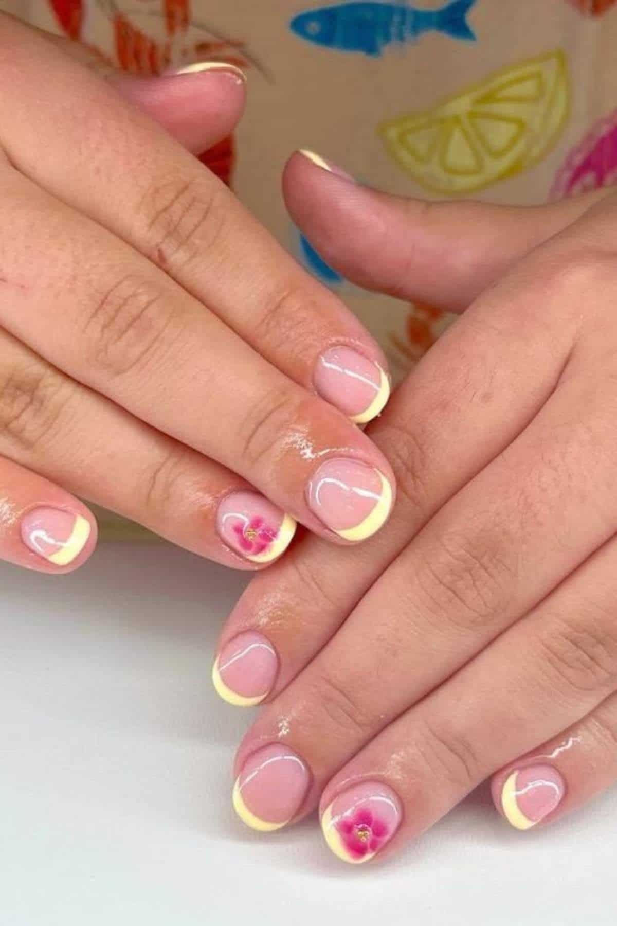 Hands with short nails featuring pale yellow French tips and small pink floral designs on two accent nails, set against a colorful shirt background.