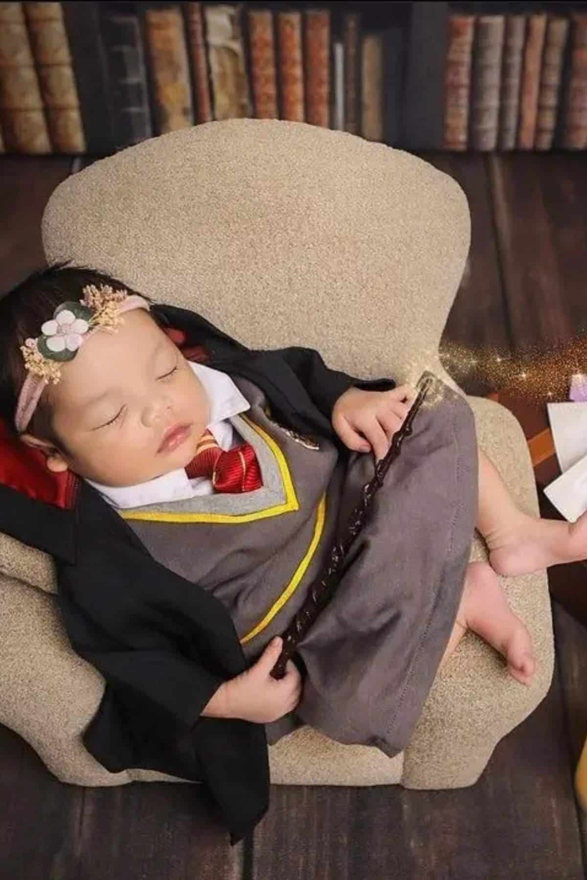 A baby dressed in a Harry Potter-inspired costume sleeps in a small armchair, holding a wand, with bookshelves in the background.