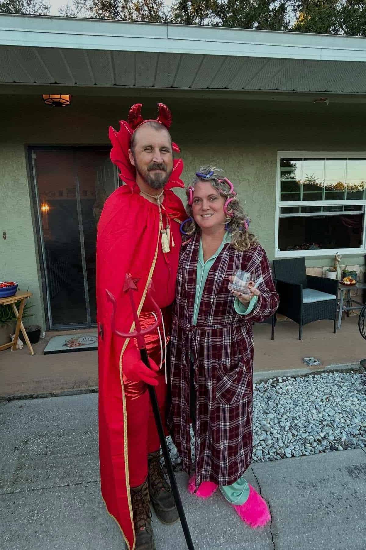 Two adults in costumes stand outside a house; one is dressed as a devil in red, the other in pajamas with curlers, holding a drink.