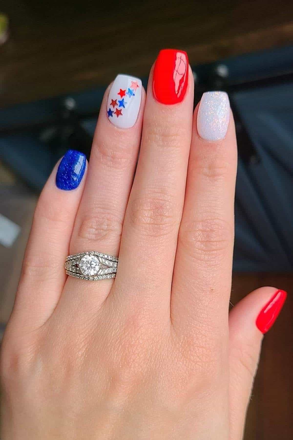 A hand with manicured nails in red, white, and blue with star designs, wearing a diamond ring on the ring finger.