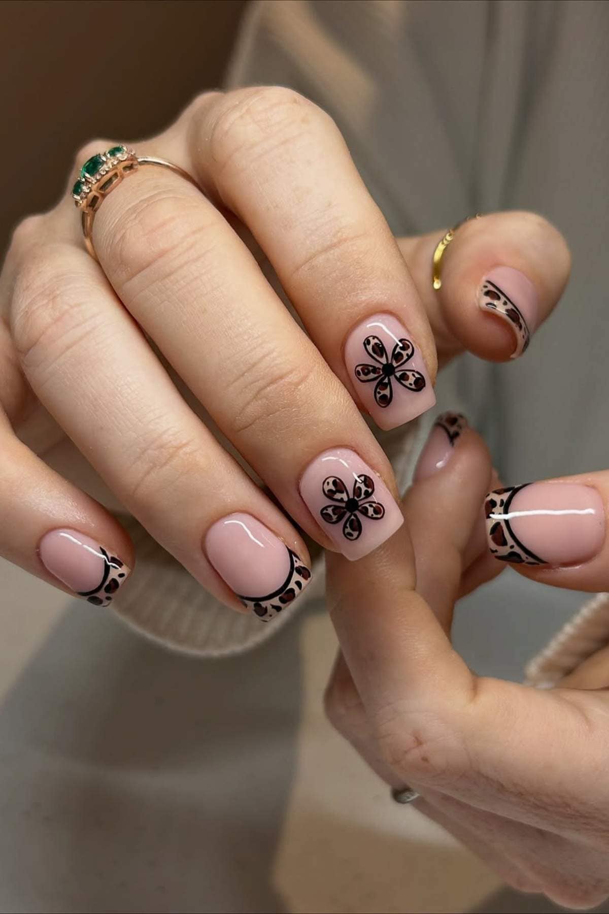 A hand with short, square-shaped nails painted in pale pink, featuring black floral designs on some nails and black patterned tips on others.