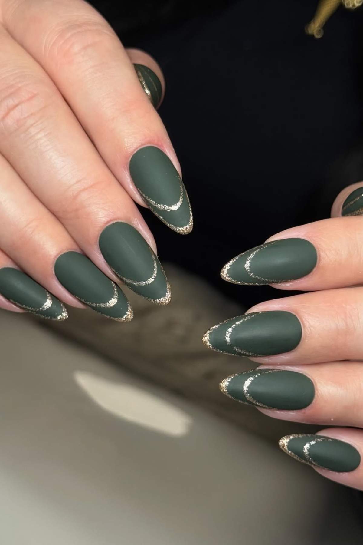 Hands with almond-shaped nails painted matte dark green, each outlined with a thin gold glitter accent near the tips.