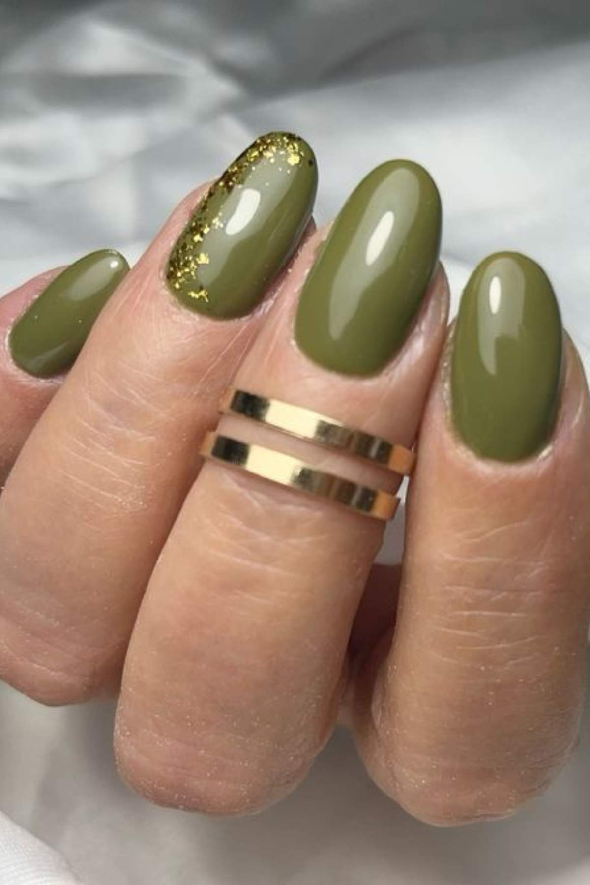 A hand with olive green oval-shaped nails, one accent nail with gold glitter at the base, and a double gold ring on the middle finger.