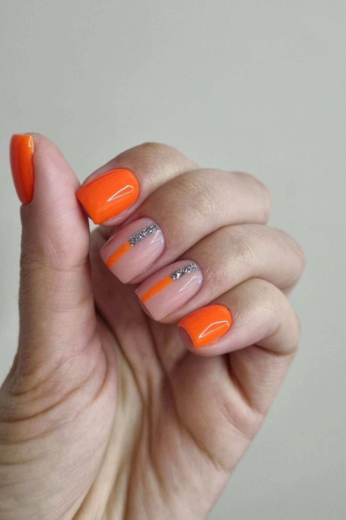 A hand with a bright orange manicure; two nails feature silver glitter accents and orange tips on a nude base.