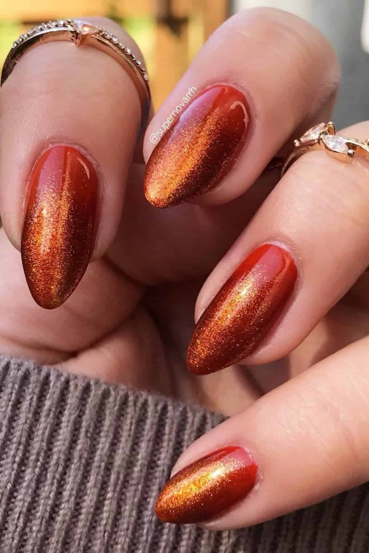A hand with almond-shaped nails painted in a shimmery, metallic red-orange polish. The person is wearing gold rings and a brown knit sleeve is partially visible.