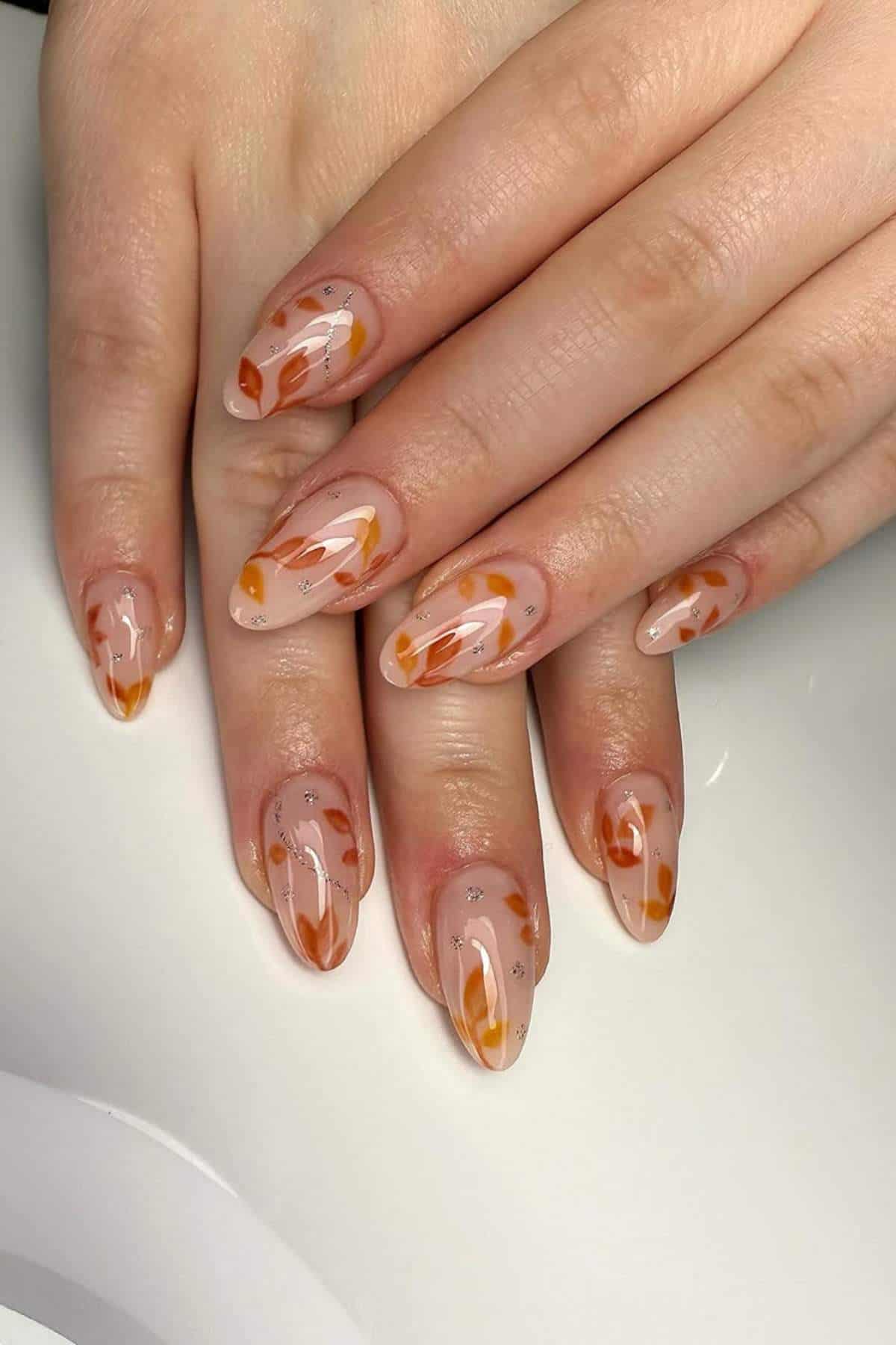 Hands with long almond-shaped nails featuring a nude base, orange and yellow swirl designs, and small silver accents.