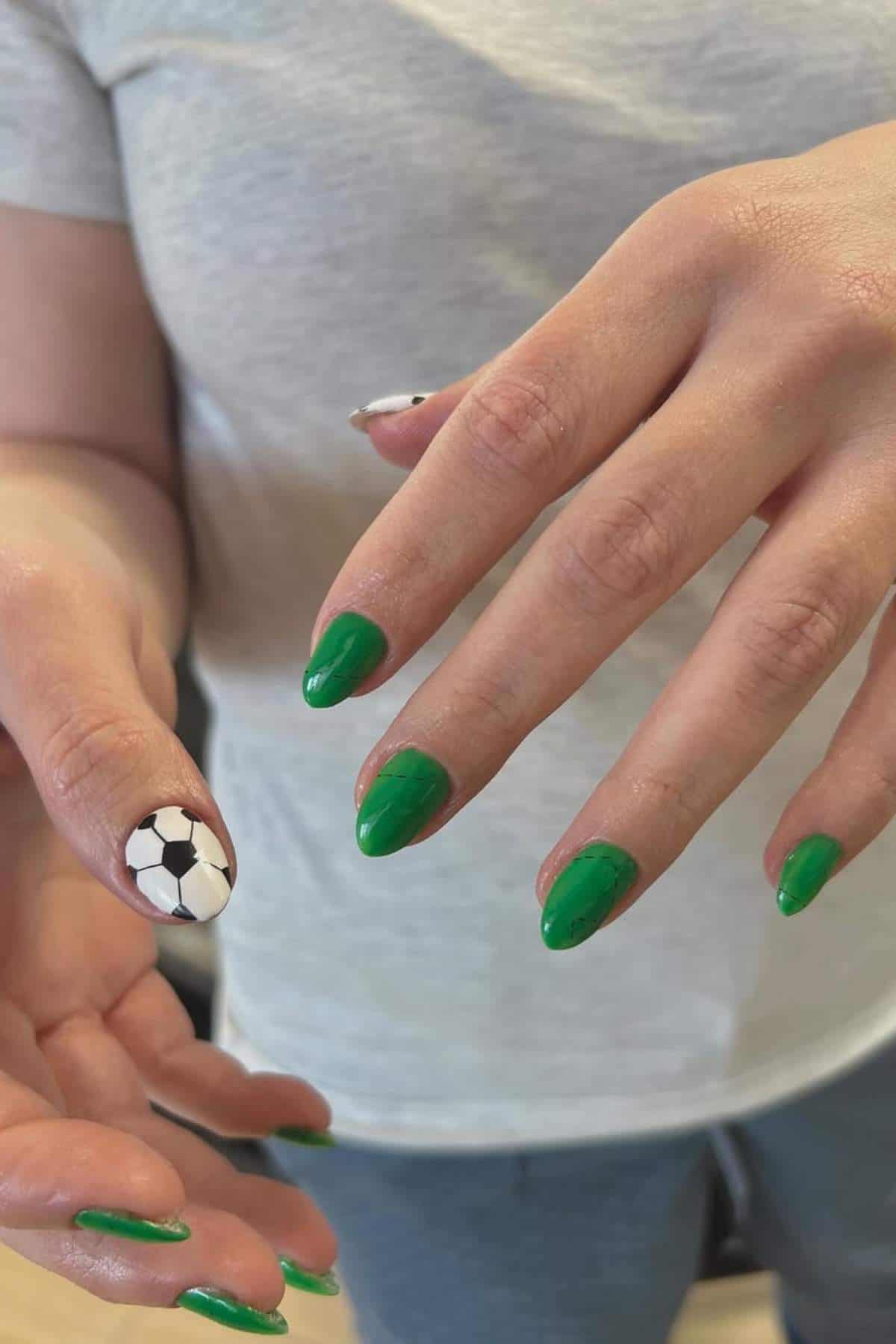 A person with green manicure and an accent nail painted like a soccer ball on the thumb, wearing a light gray shirt.