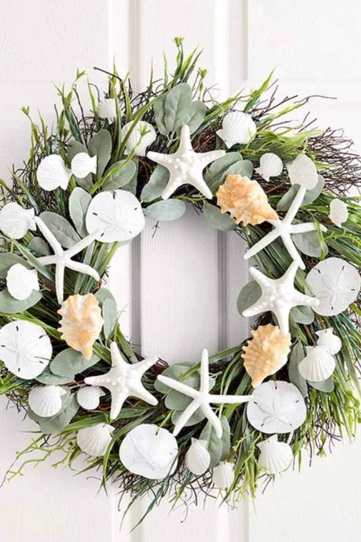 A wreath made of green foliage, white starfish, sand dollars, and seashells, hanging on a white door.