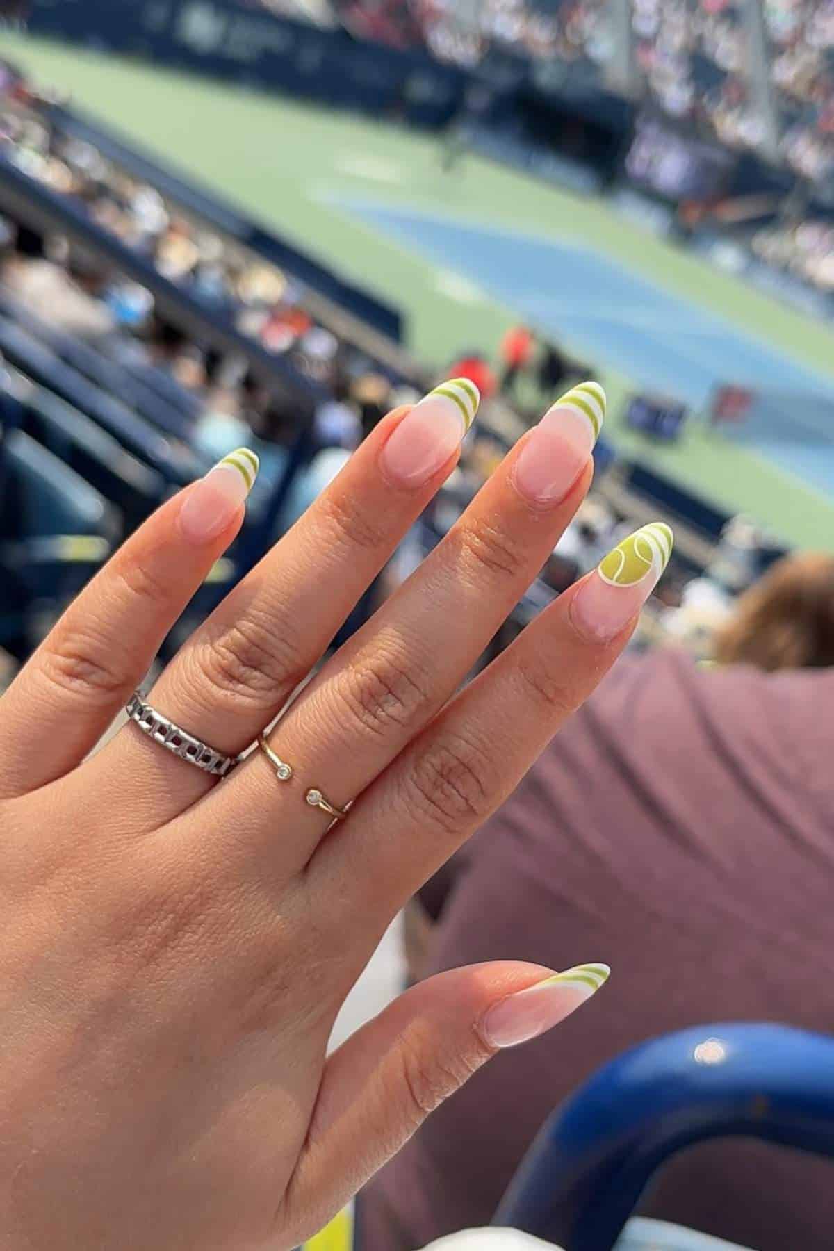 A hand with long, almond-shaped nails featuring green and white swirl designs is held up at an outdoor tennis stadium.