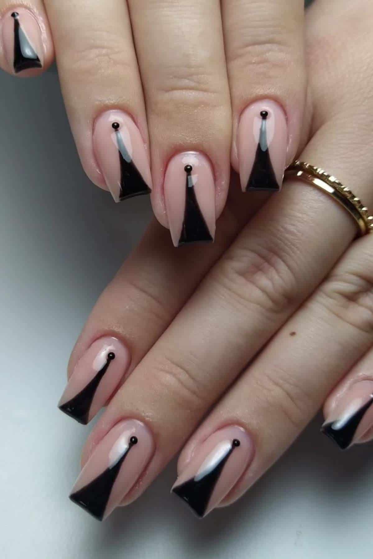 Close-up of hands with nude nails featuring black geometric triangle designs and small black dots at the base, paired with gold rings.