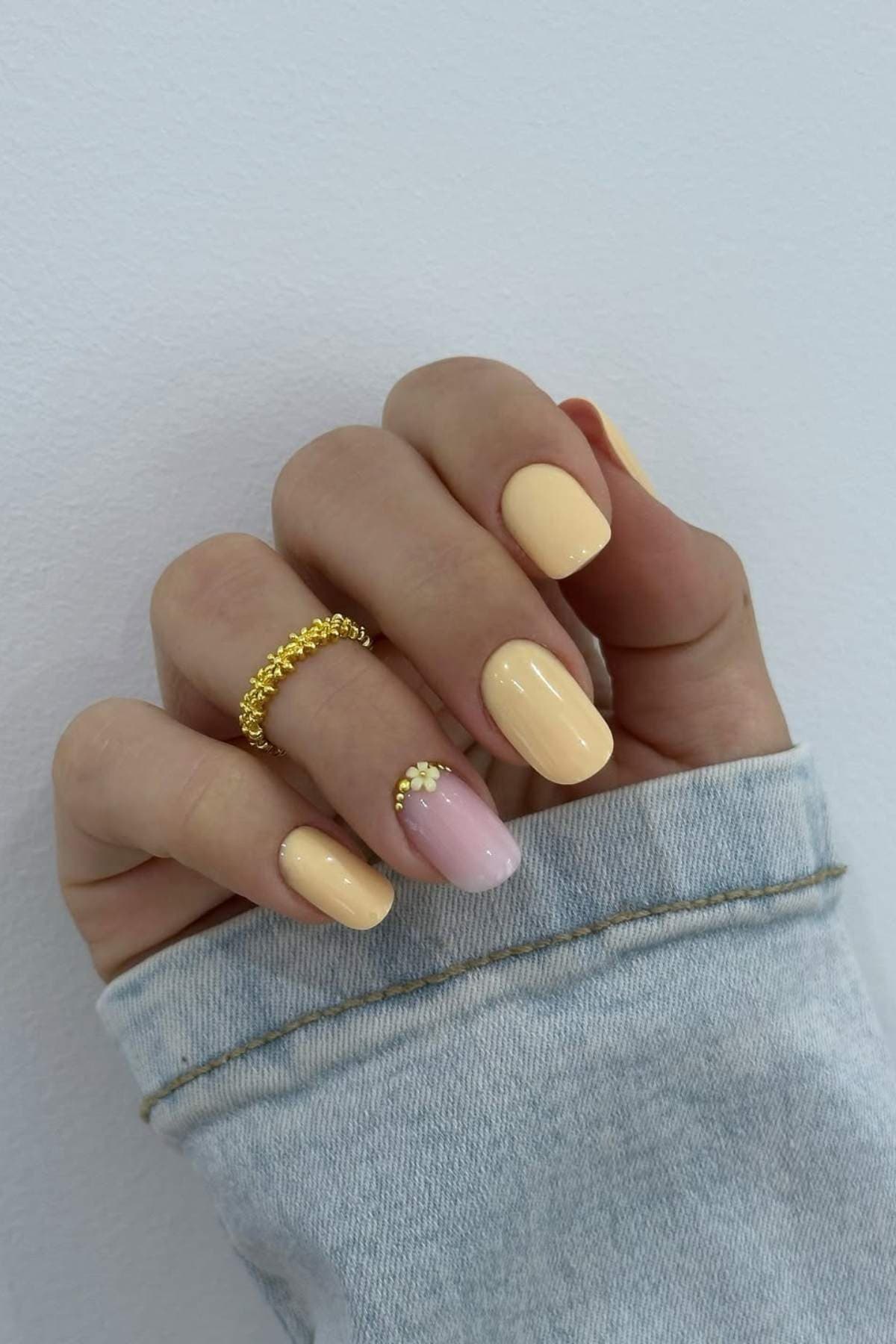 A hand with short, almond-shaped nails painted in pale yellow, except for the ring finger, which is light pink with small yellow studs near the cuticle. The person is wearing gold rings.