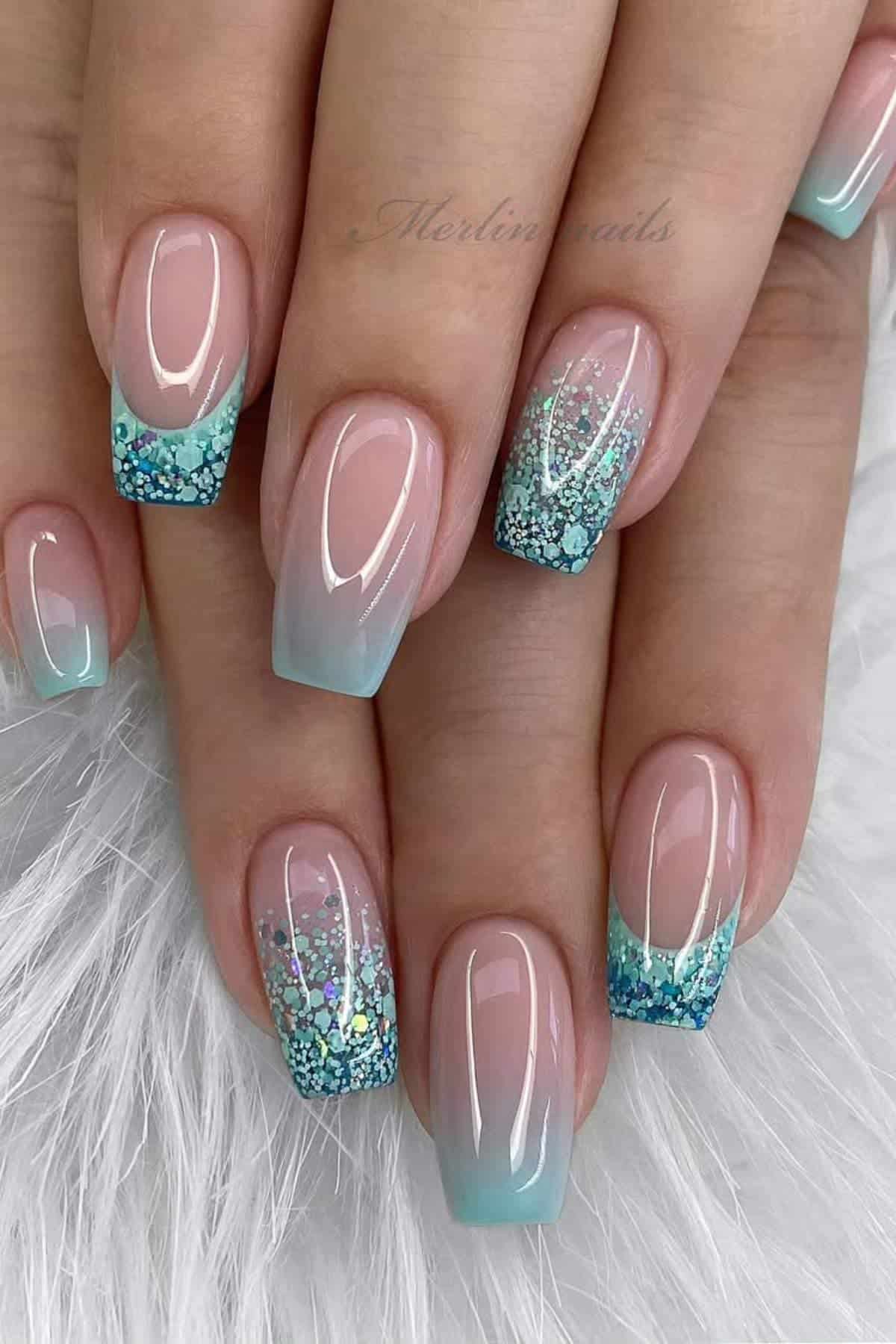 Close-up of hands with square-shaped nails featuring a light blue ombre manicure and blue glitter accents on the tips, displayed on a white textured surface.