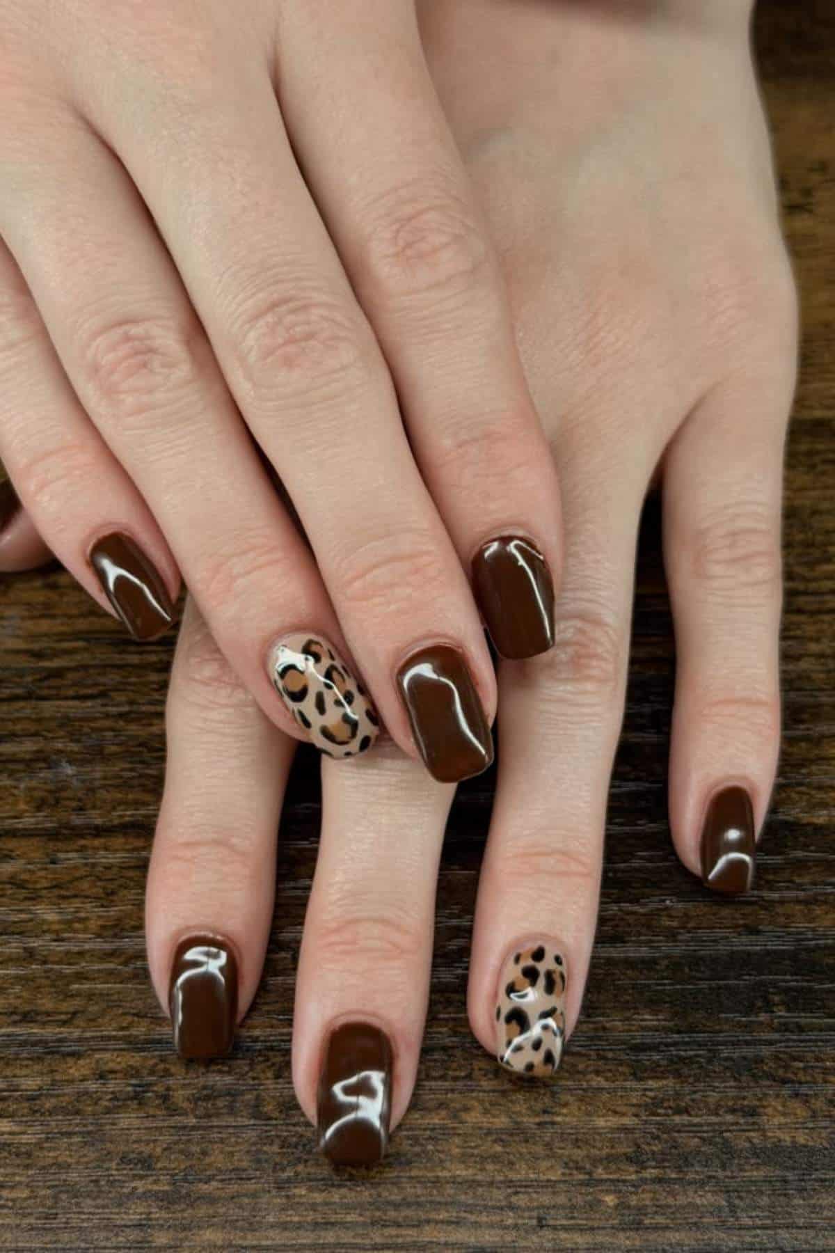 Hands with brown square-shaped nails; two accent nails feature a leopard print design. Nails rest on a wooden surface.