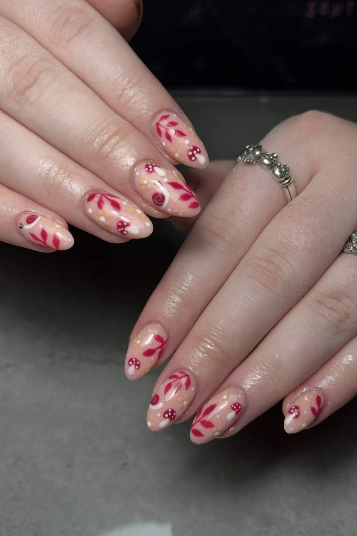 Hands with almond-shaped nails featuring a nude base, red leafy designs, small red dots, and white accents; person wearing a silver ring with small beads.
