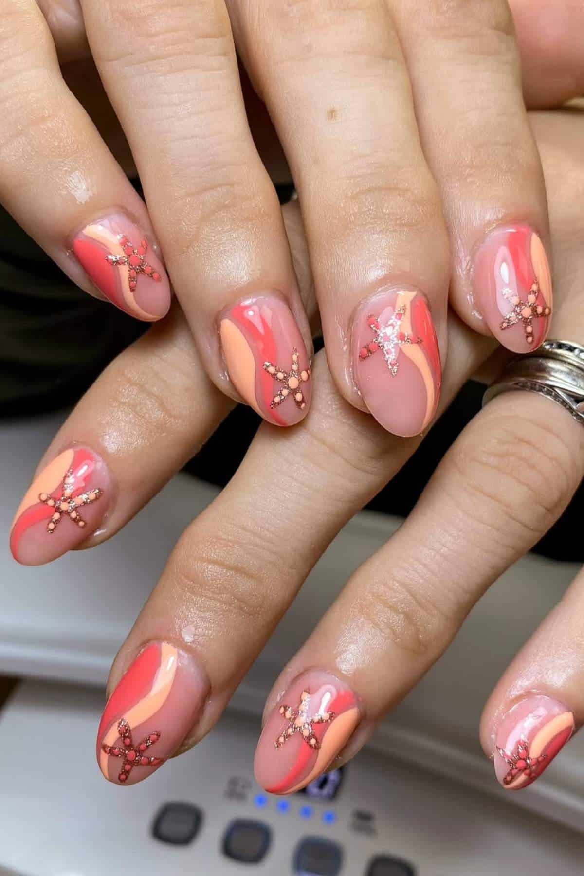 Hands with almond-shaped nails featuring a pink and peach abstract design, embellished with rhinestone star patterns on each nail.