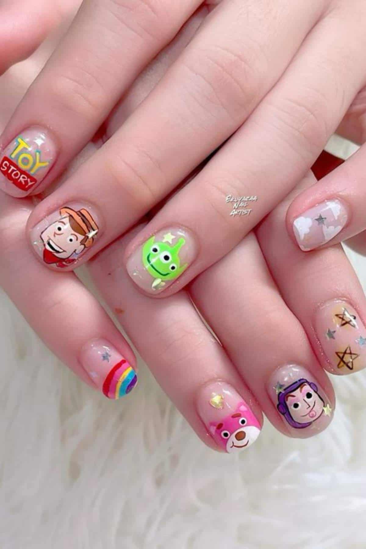 Hands with manicured nails featuring Toy Story-themed nail art, including characters and symbols from the animated movie on a light, natural base.