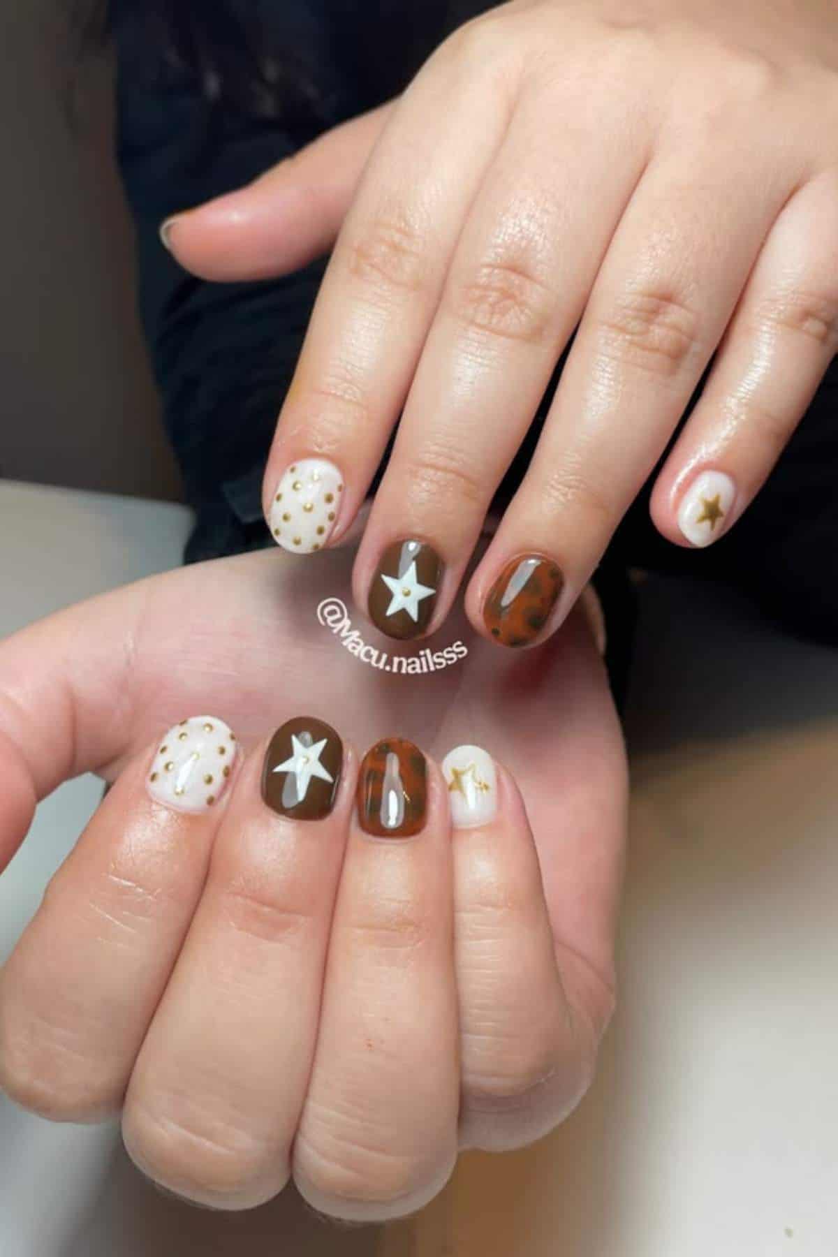 Hands with short nails painted in brown and white tones, featuring star designs and small polka dots on some nails.
