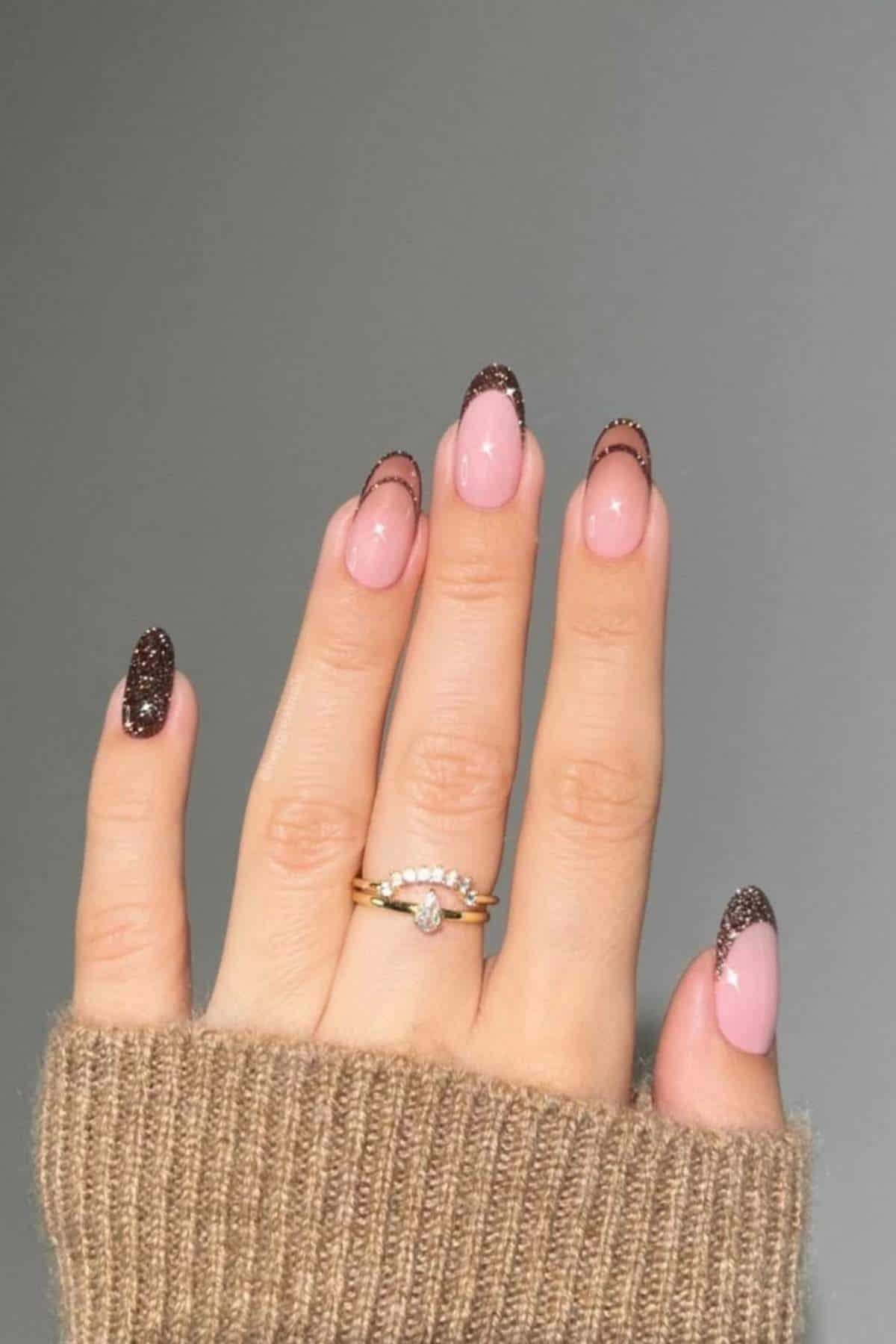 A hand with almond-shaped nails featuring pink polish and dark brown glittery tips, wearing two gold rings, and partially covered by a brown knit sleeve.
