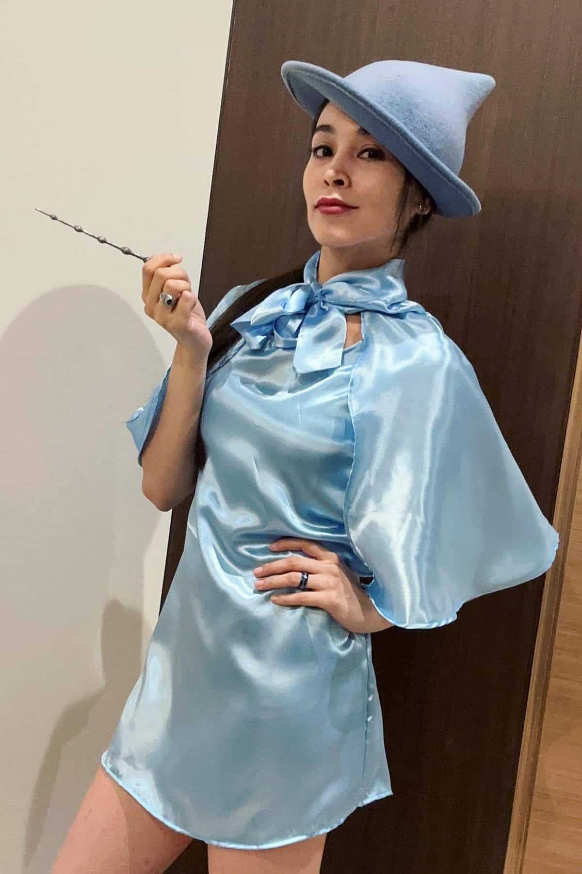 Person wearing a light blue shiny dress and matching hat, holding a wand, standing indoors in front of a wooden door.