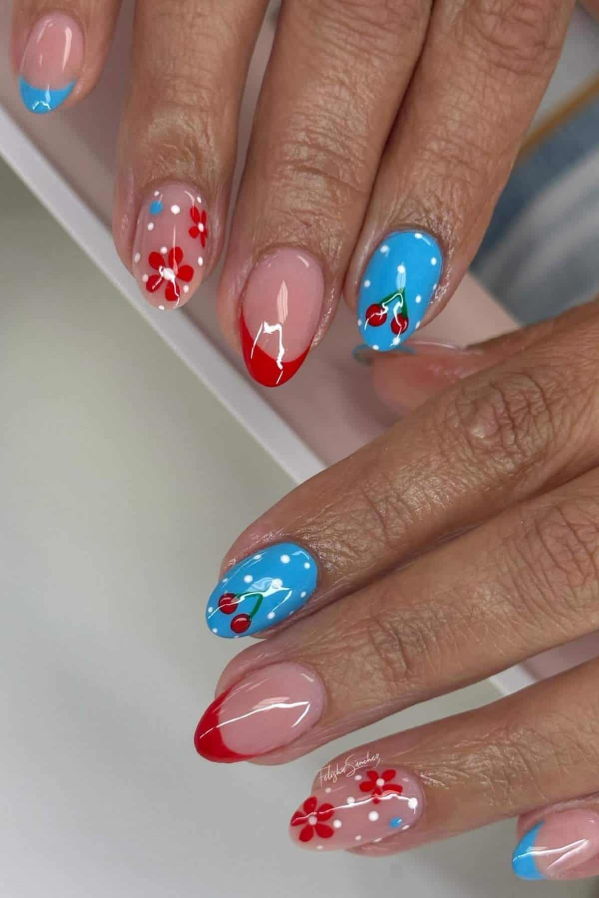 A hand with almond-shaped nails featuring red French tips, blue accents, and nail art with red flowers, cherries, and white dots.