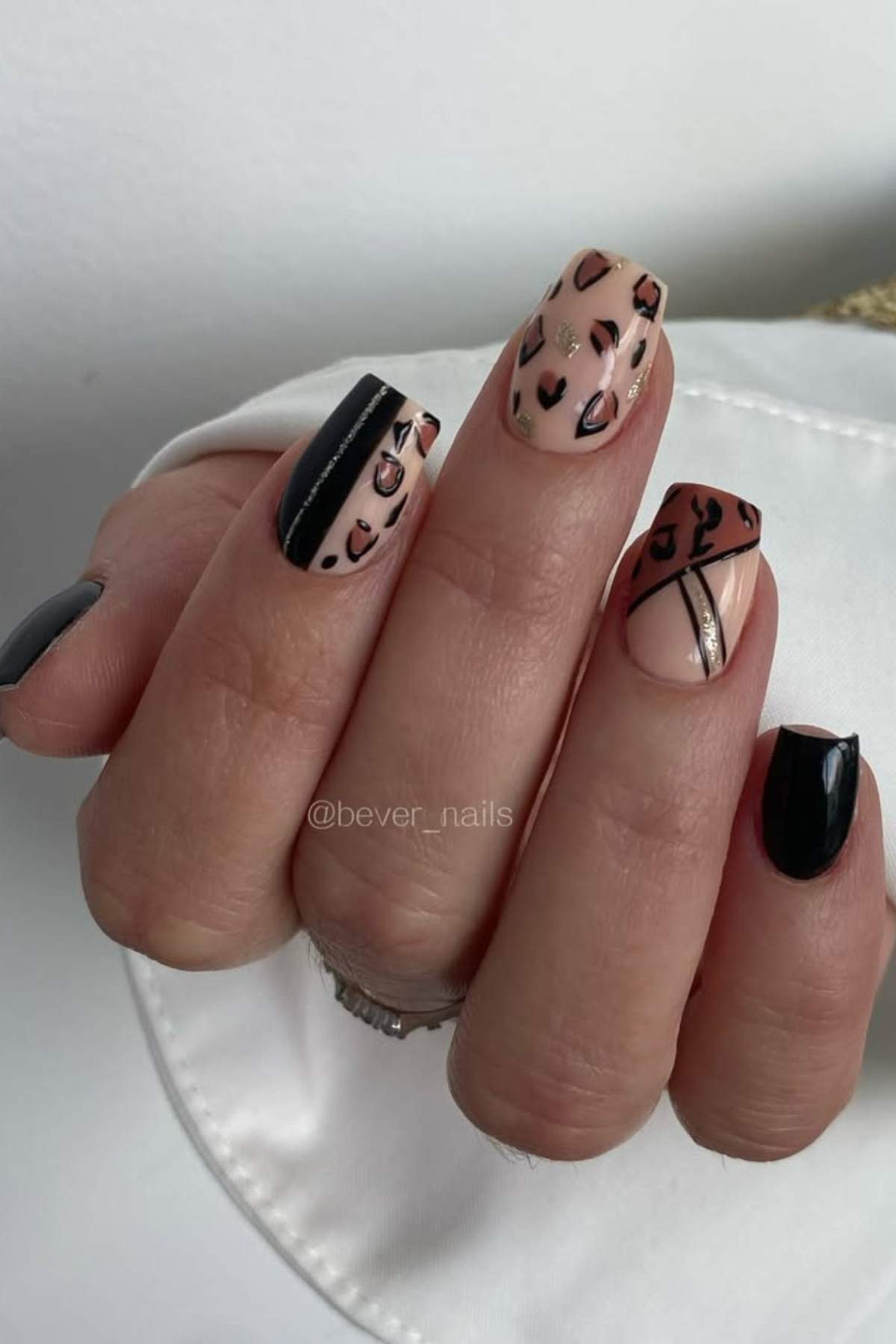 A hand with short square nails featuring black, beige, and brown nail art, including leopard print, black stripes, and geometric designs.