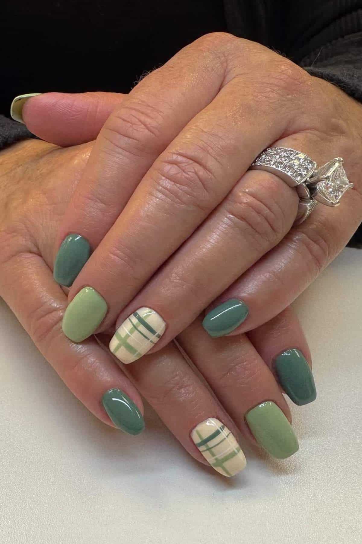 Hands with manicured nails in various shades of green and ring finger accent nails featuring a green plaid design. Person is wearing two diamond rings.