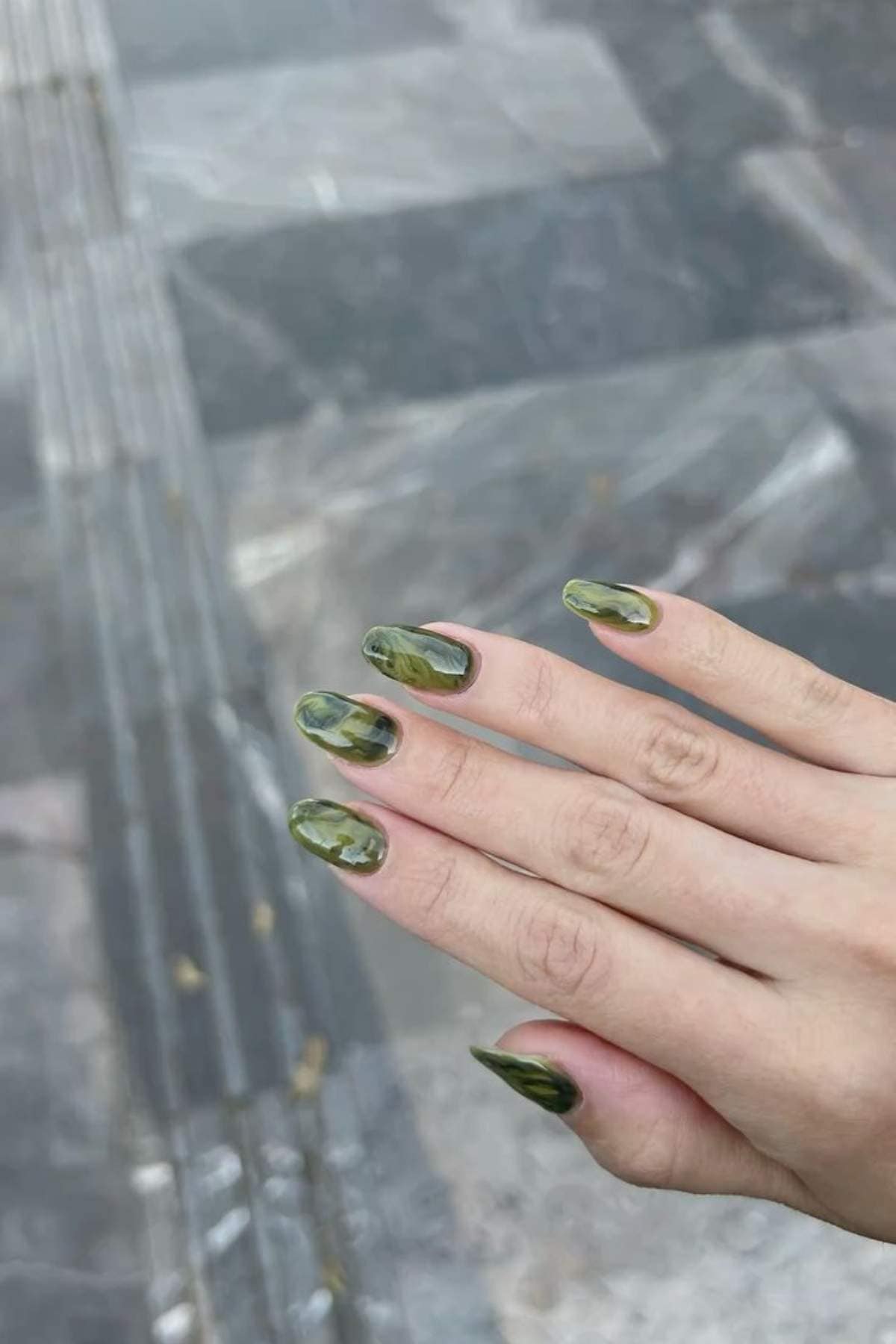 A hand with long, almond-shaped nails painted in a green and black marble pattern is shown against a tiled outdoor surface.