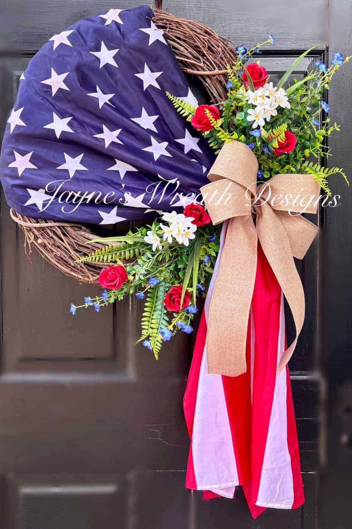 Wreath made of grapevine with American flag fabric, red and white flowers, greenery, and a burlap bow on a black door.
