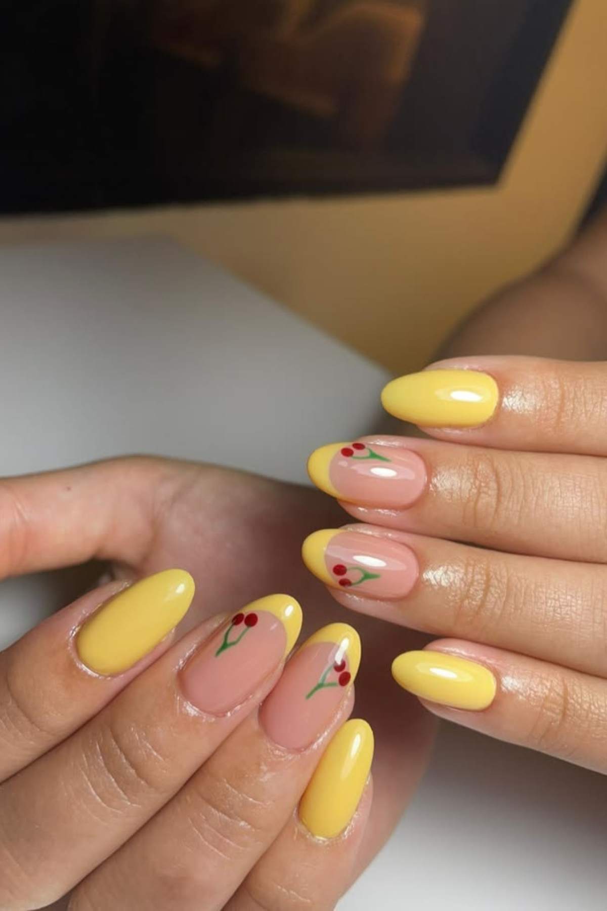Close-up of hands with almond-shaped nails painted yellow and nude, featuring small red cherry designs on some accent nails.
