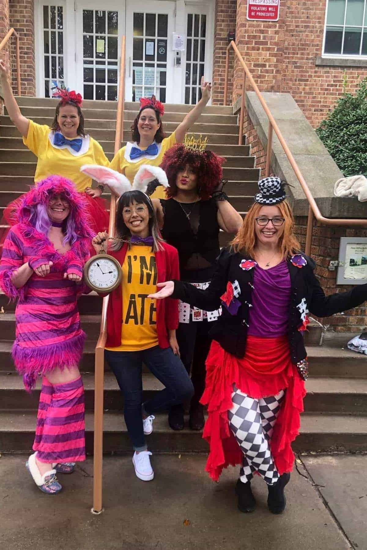 Six adults in colorful costumes, including Alice in Wonderland characters, pose on outdoor steps in front of a brick building.