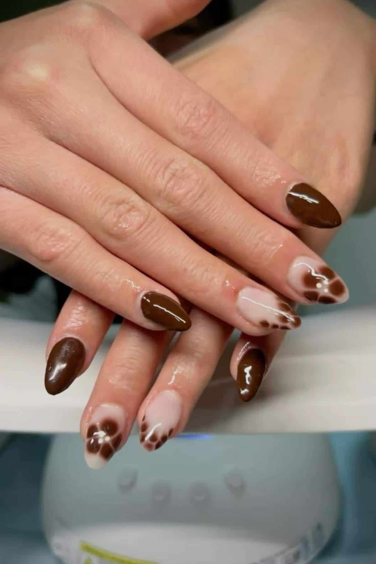 Hands with almond-shaped nails featuring a brown and nude cow print nail art design, alternating with solid brown nails.