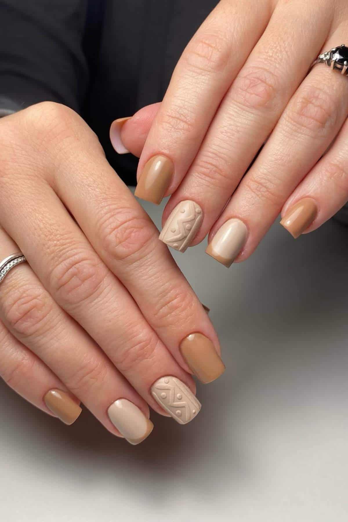 Close-up of hands with short, square-shaped nails painted in nude and tan shades; one accent nail features a textured geometric pattern.
