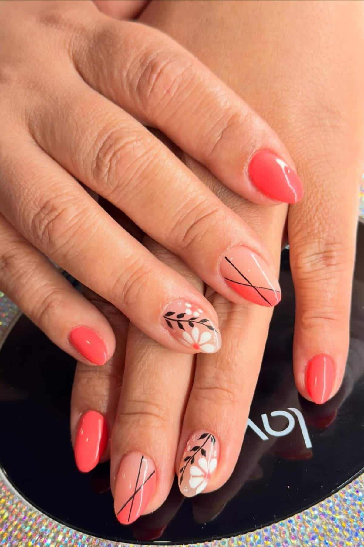 Hands with coral pink manicured nails; two nails feature floral and line designs in black and white on a nude base.